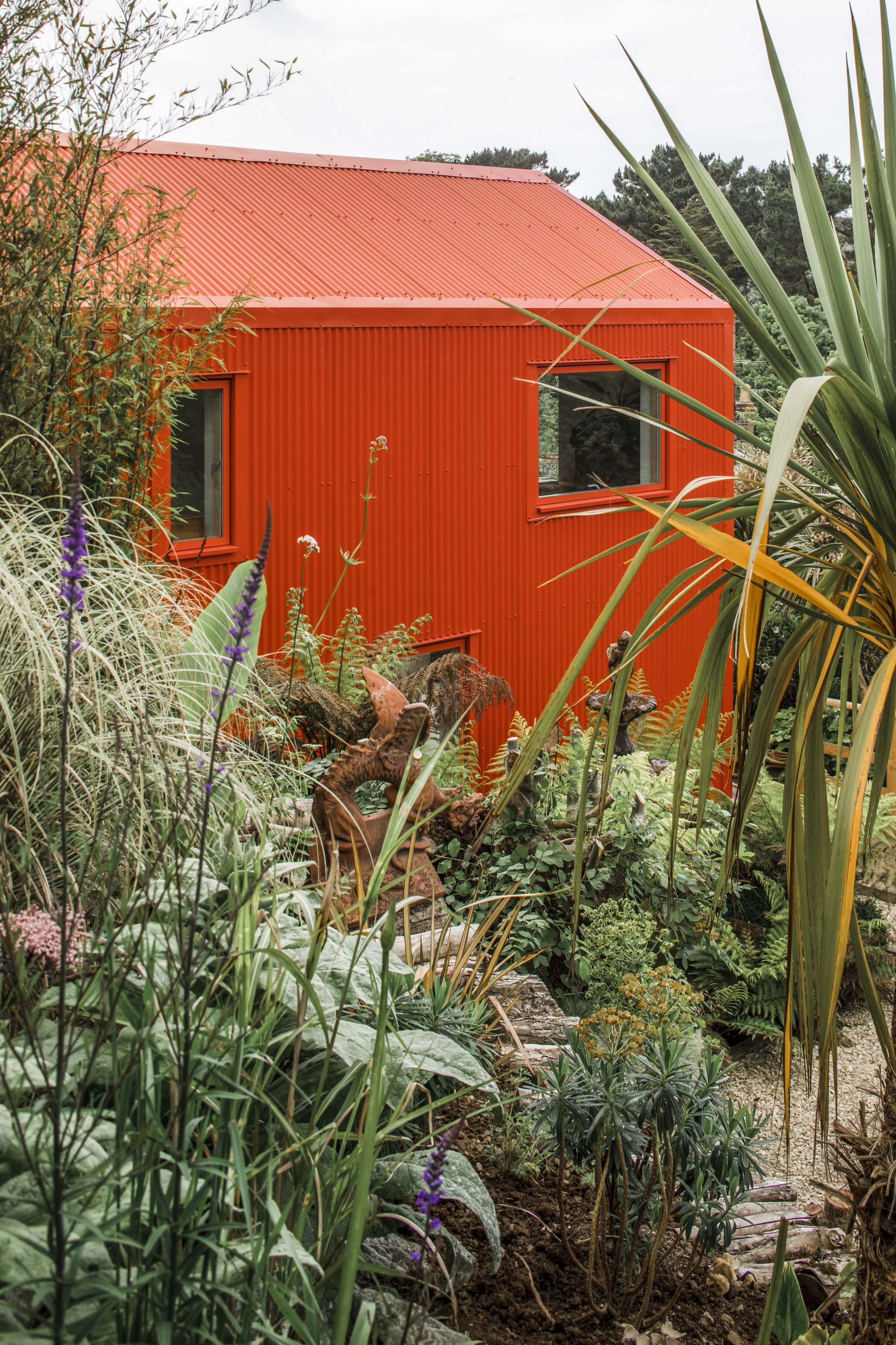 A bold, corrugated structure set within dense planting, its vibrant red exterior creating a striking contrast against lush greenery. The building feels both embedded in and distinct from its landscape, balancing playfulness with functional clarity.