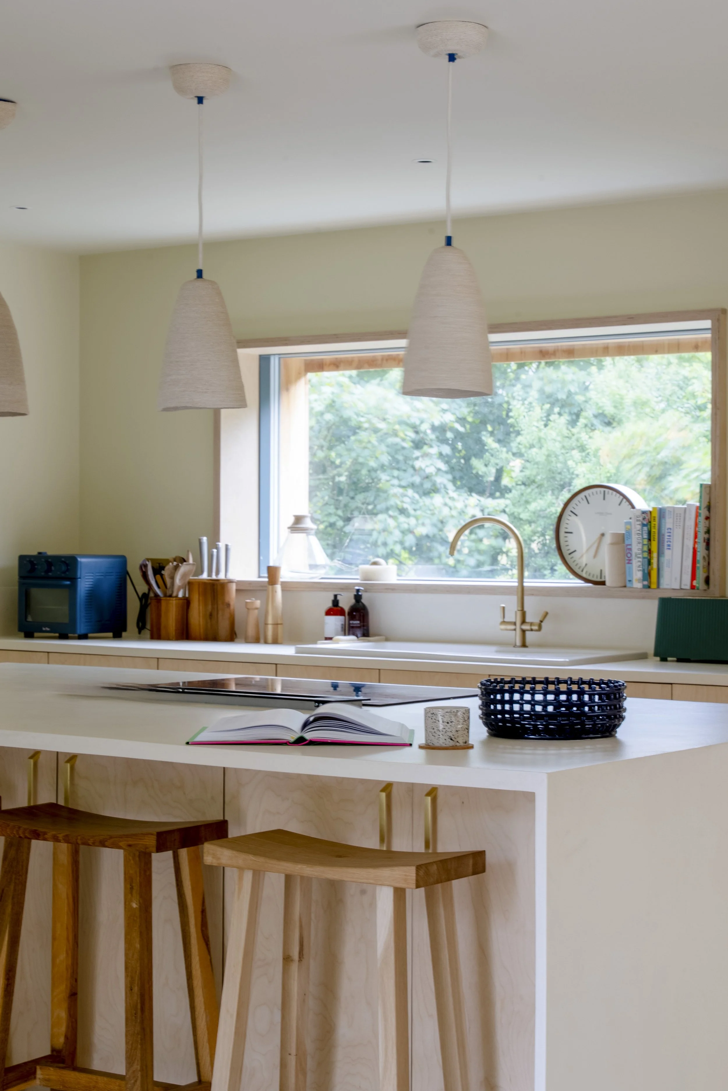 Light-filled and practical, this kitchen balances clean lines with handcrafted details. Pale timber joinery, woven pendant lighting, and simple shelving create a welcoming environment designed for daily use, with views out to greenery providing a con