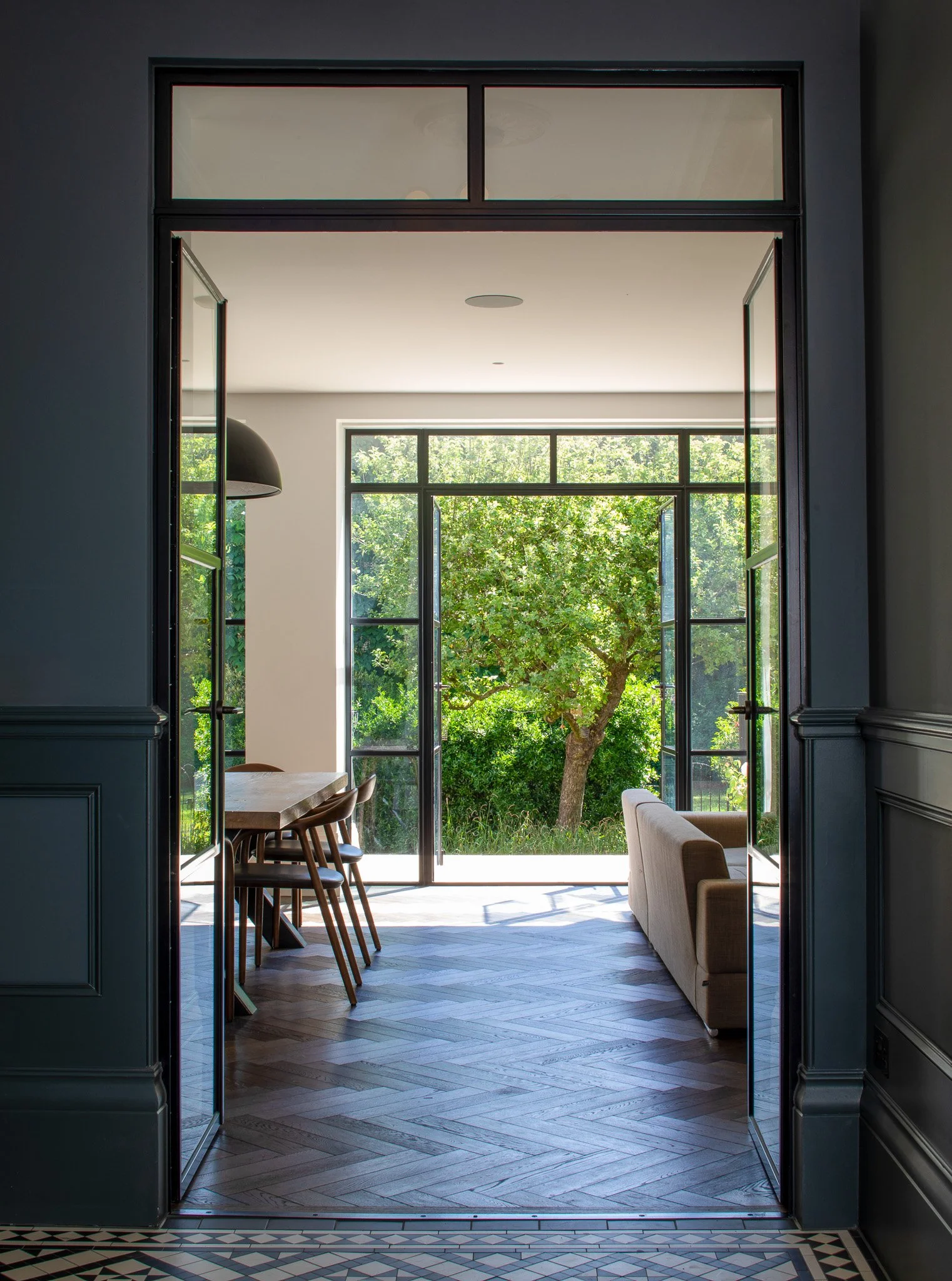A carefully framed view through full-height glazed doors reveals a seamless connection between interior living spaces and the garden beyond. Dark detailing and herringbone timber flooring create a sense of depth, while natural light draws the eye out