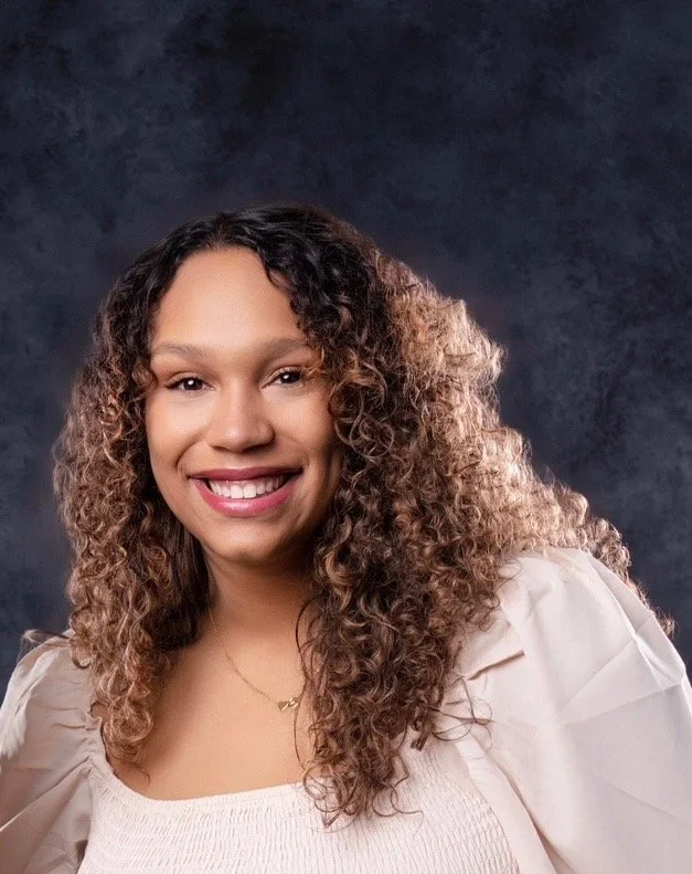 A woman with curly brown hair smiling at the camera, wearing a white top and a delicate necklace, against a dark textured background.