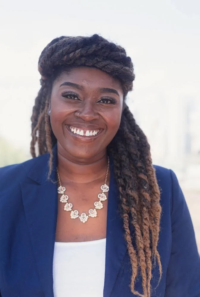 A smiling woman with dark skin, wearing a navy blazer, white top, and a silver necklace, outdoors with a blurred background.
