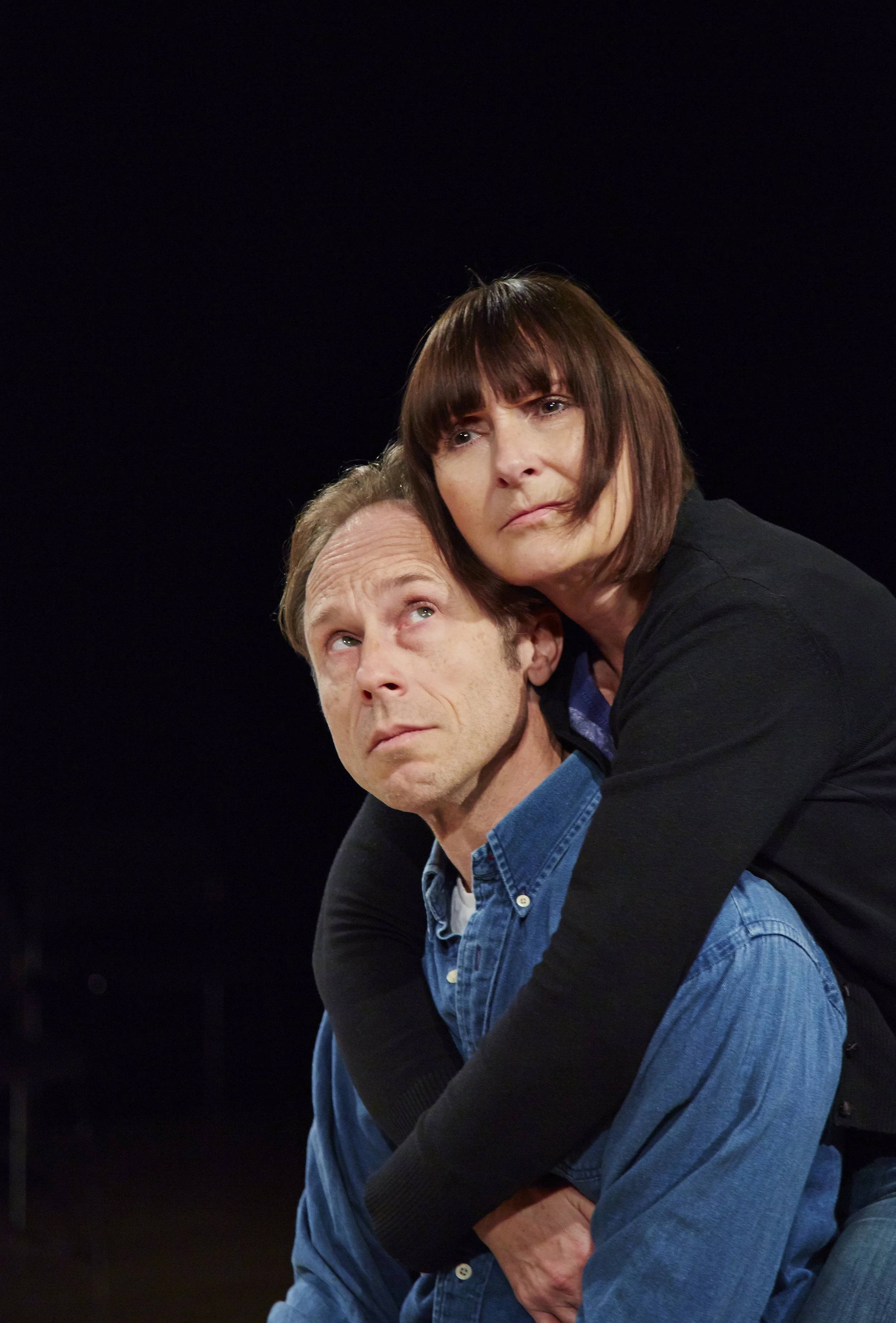 A man with short, light brown hair and a woman with dark, straight hair with bangs are engaged in a dramatic scene. The woman is leaning over the man, who is sitting and looking up at her with a serious expression. Both are wearing casual clothing, and the background is dark.