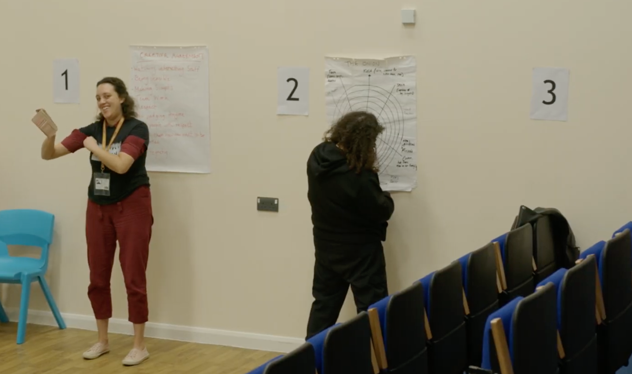 Two women standing near wall posters in a conference room. One woman is smiling, holding a card, and standing near a blue chair. The other woman, facing the wall, has curly hair and is wearing a black hoodie. There are numbered signs on the wall and rows of blue and black chairs in the foreground.