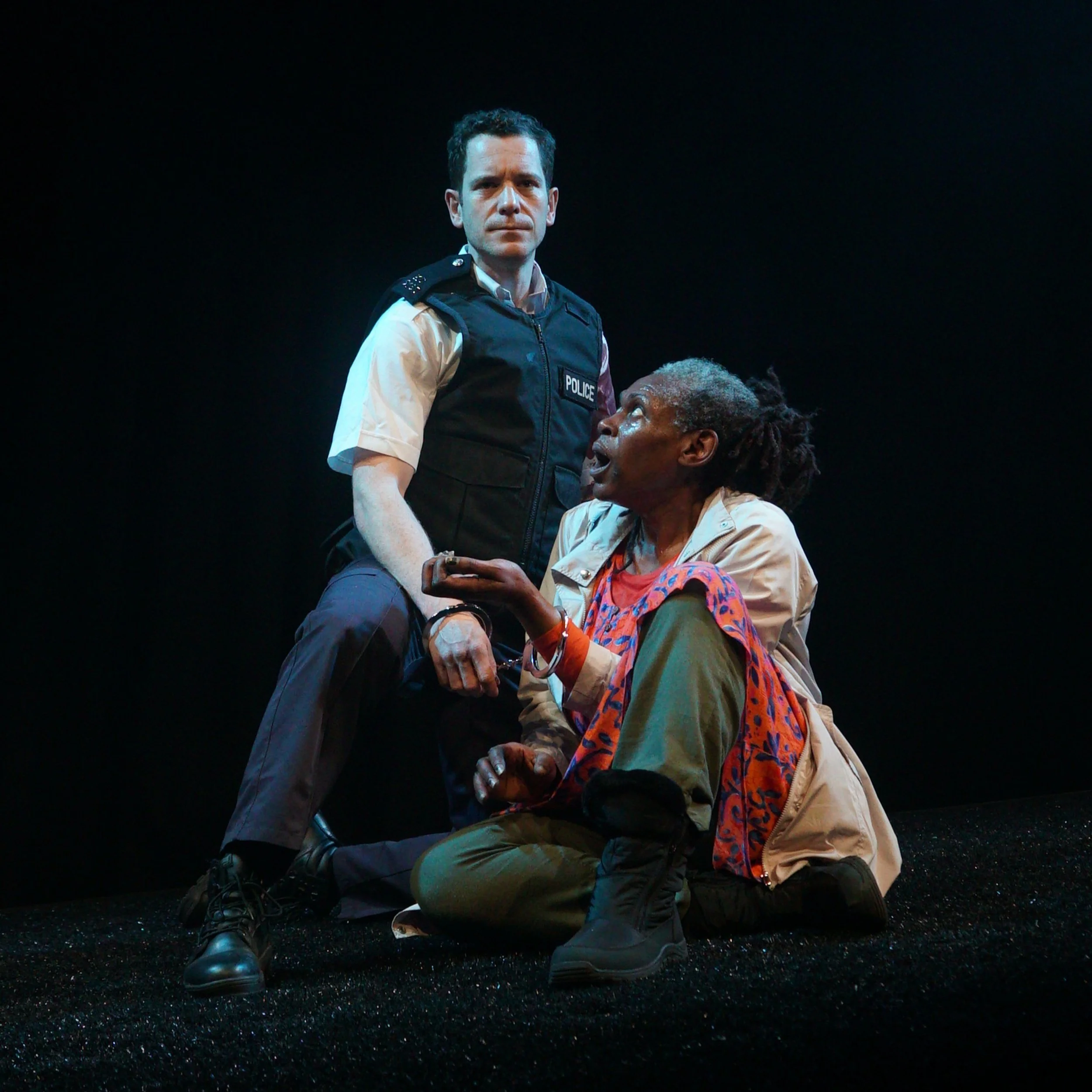 The image depicts a scene with two actors on stage. One is a man in a police uniform standing, looking serious. The other is a woman kneeling on the ground, appearing distressed and speaking to the man.