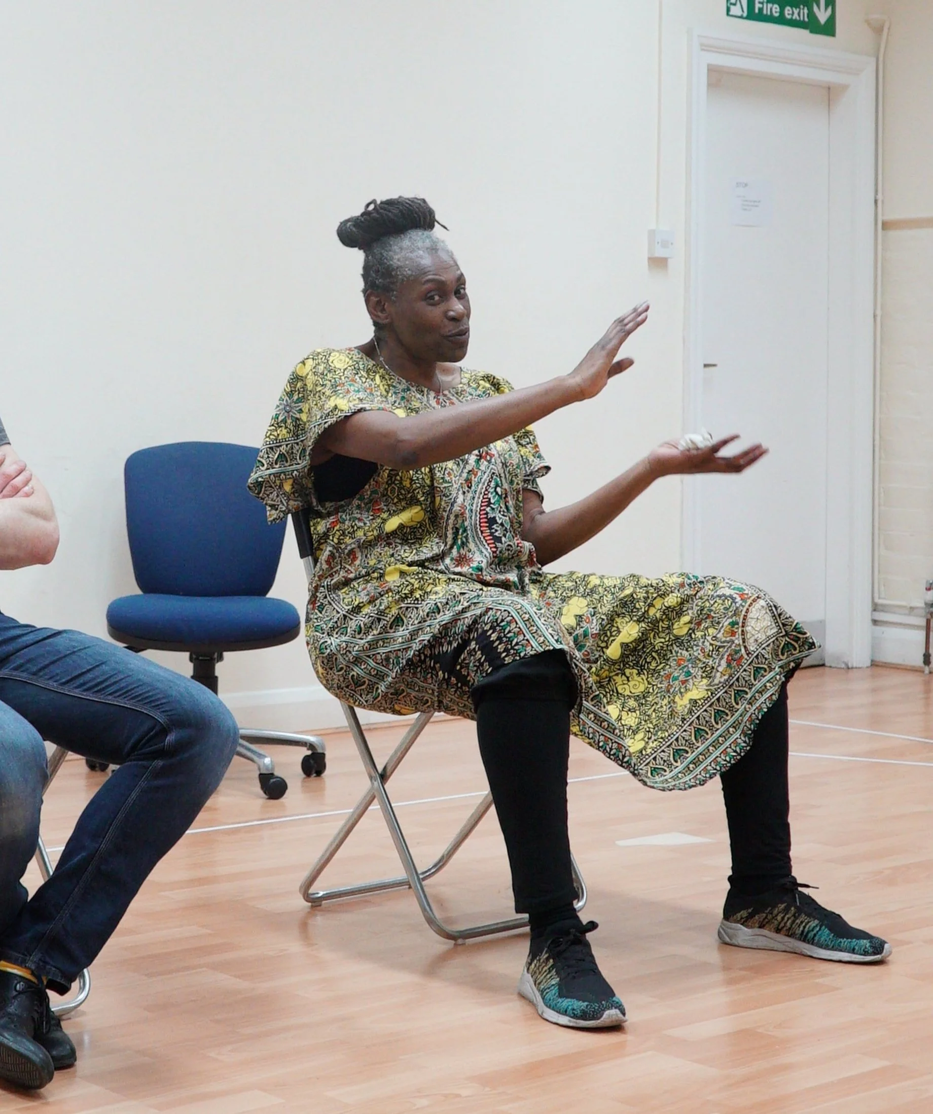 A woman with gray hair styled in an updo, wearing a patterned dress, sitting on a chair, gesturing with her hands in a room with a hardwood floor, a blue chair, and a white wall with a fire exit sign.
