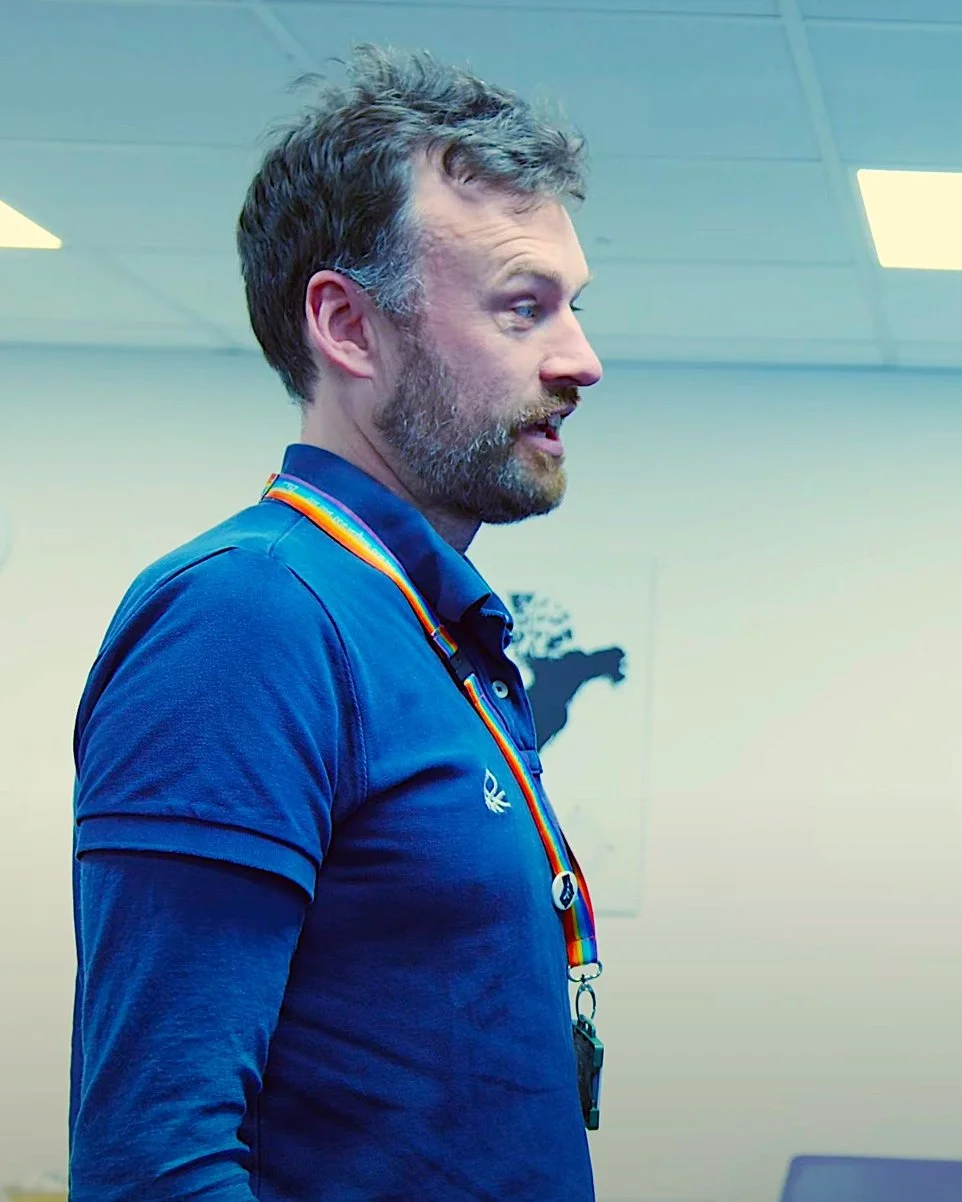 A man with a beard and short hair wearing a blue polo shirt with a rainbow-colored lanyard around his neck, standing indoors in front of a plain wall with a partial map silhouette.