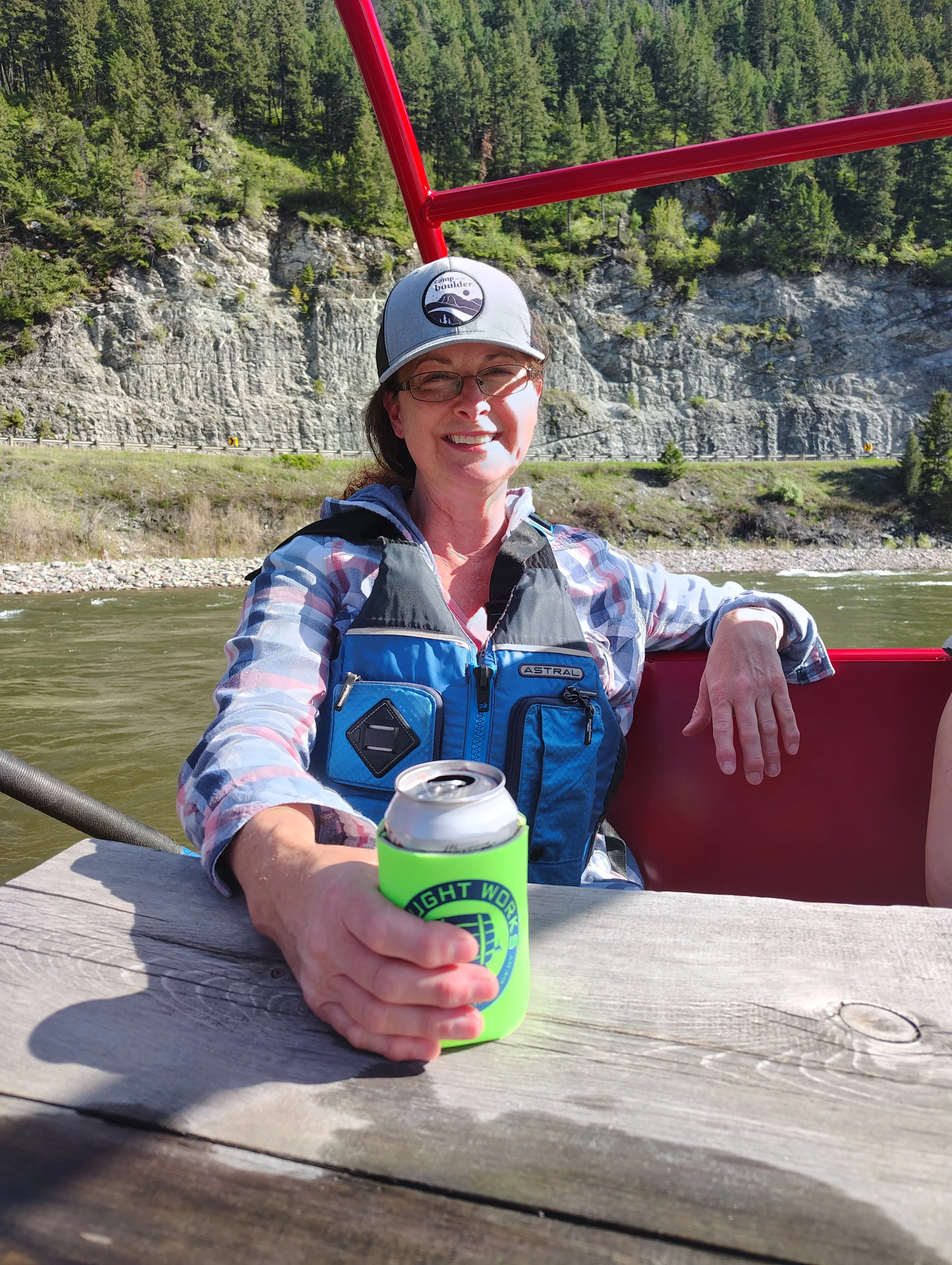 A woman smiling and sitting on a boat, holding a can of beer, with a river and forested cliffs in the background.