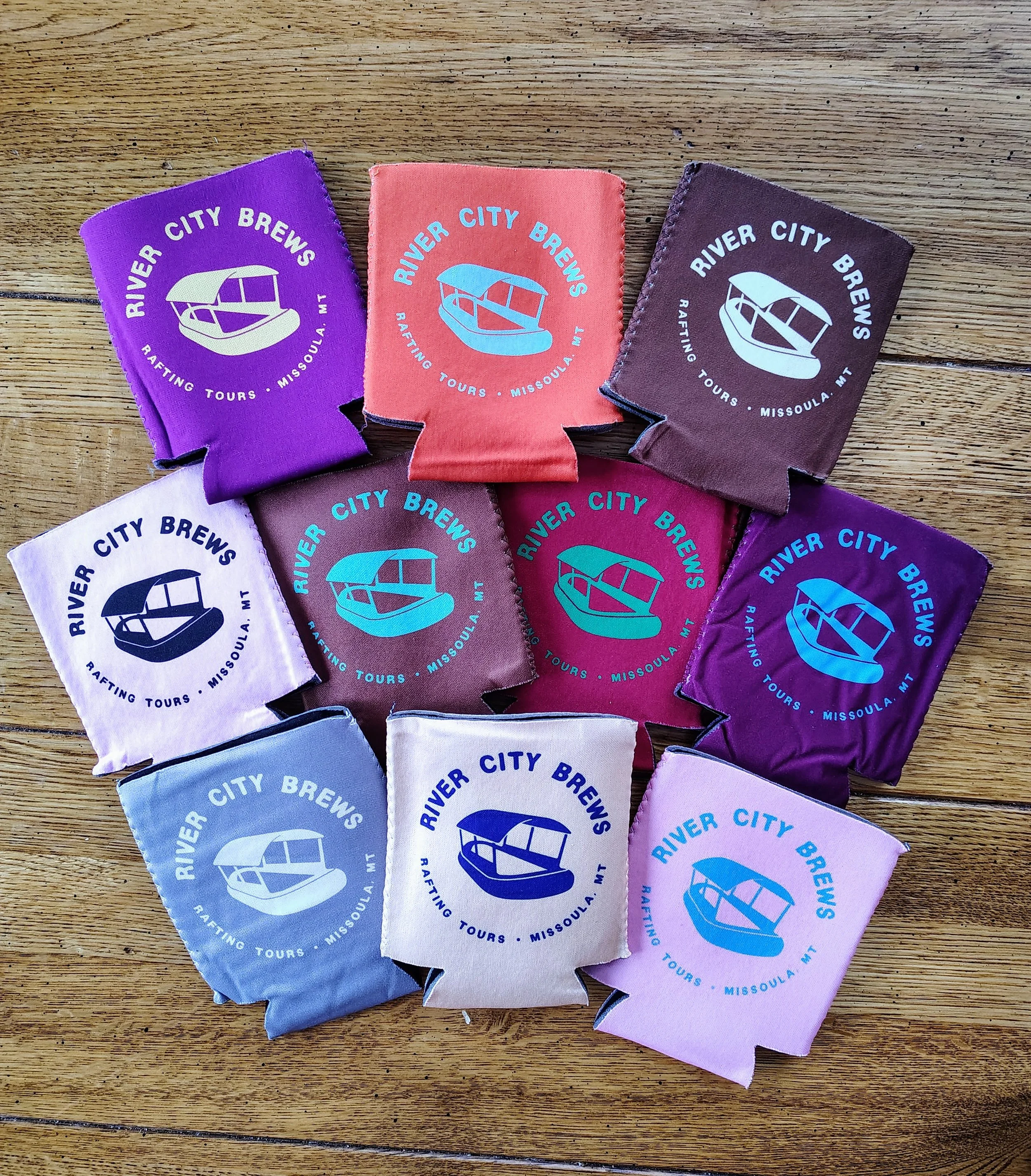 Various colored fabric drink koozies with the logo and text 'River City Brews, Rafting Tours, Missoula, MT' laid out on a wooden surface.