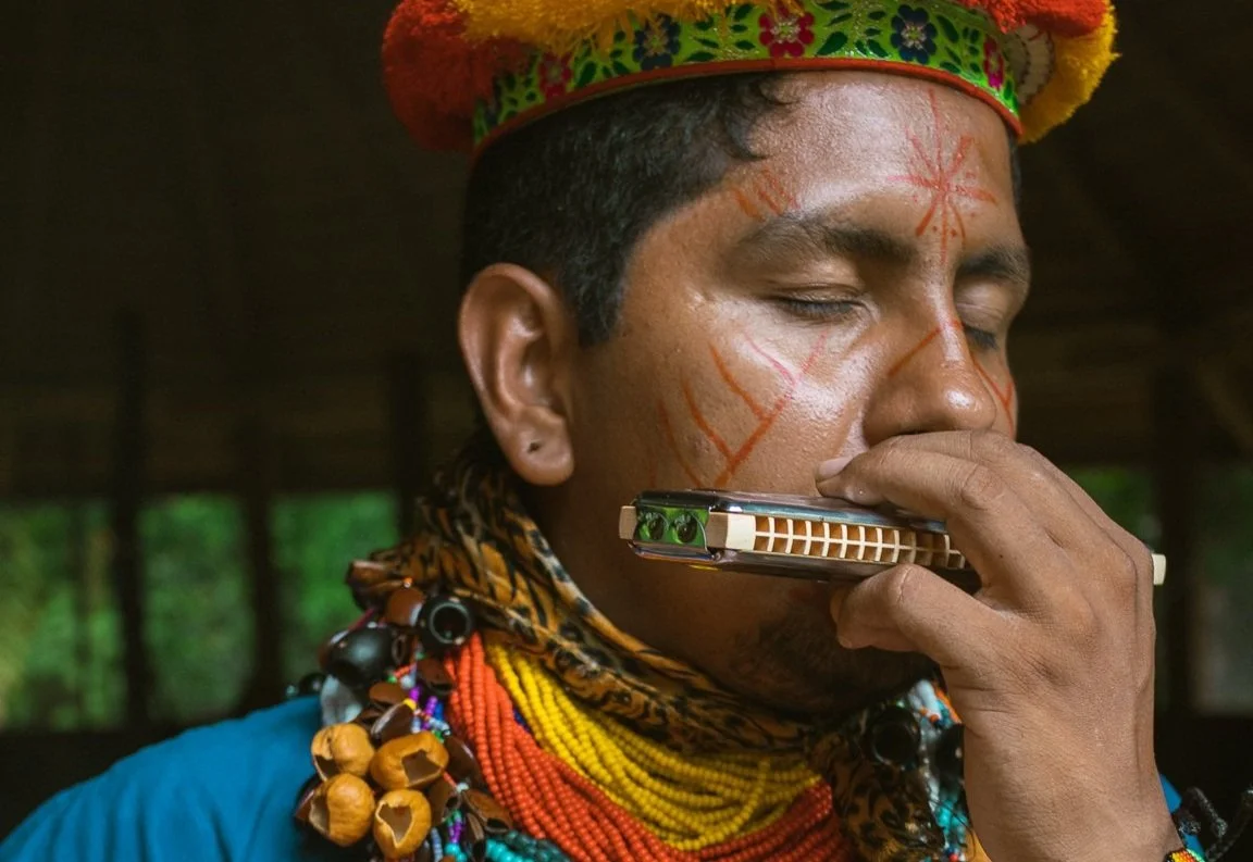 An indigenous man from an Amazon tribe wearing traditional jewelry and a colorful headdress, playing a harmonica with his eyes closed.