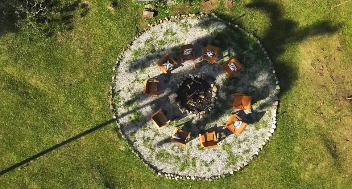 A circular fire pit surrounded by ten wooden chairs on a stone and gravel surface, with grass and trees around the area.