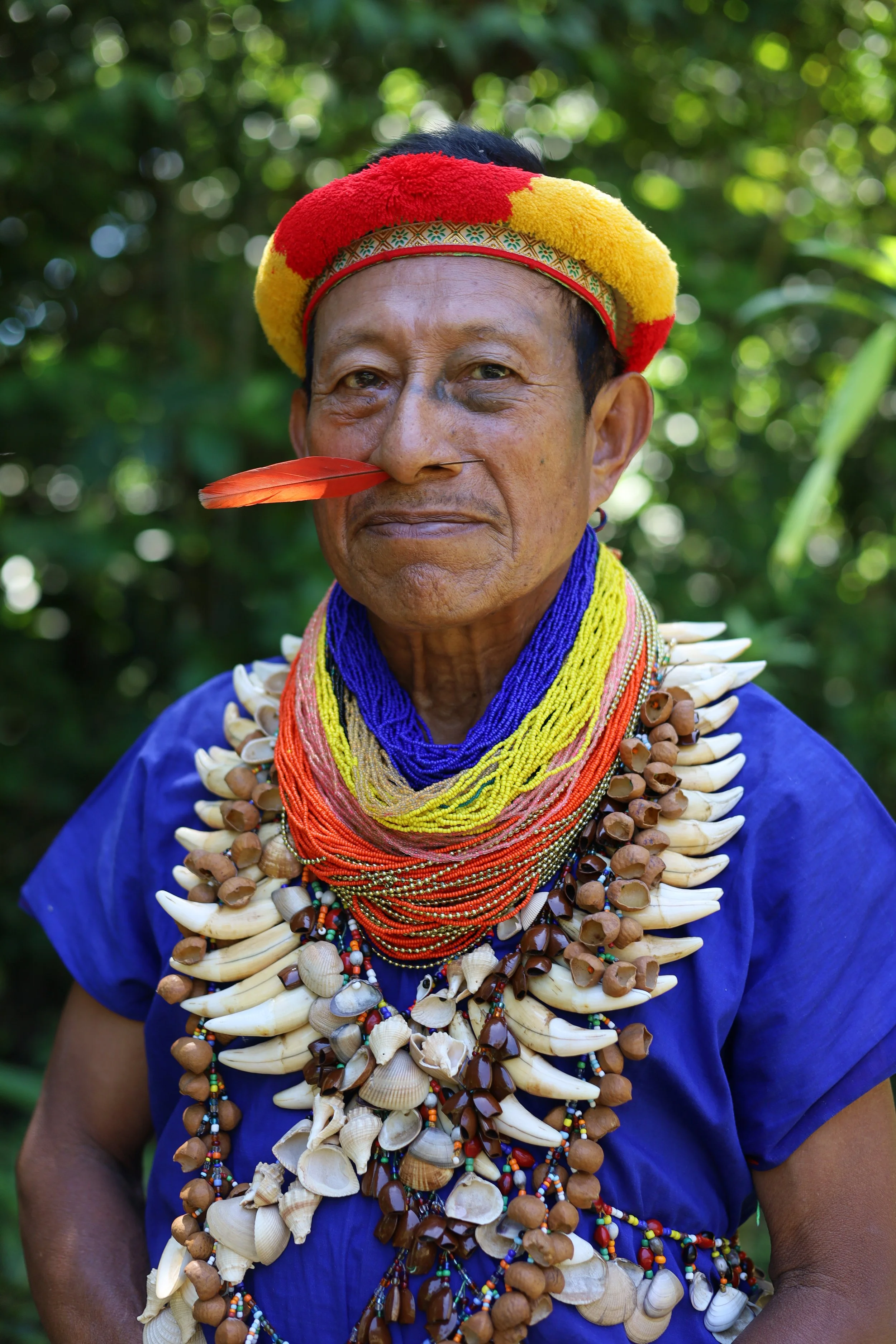 A woman wearing traditional attire with multiple colorful beaded necklaces and shell decorations. She has a red, yellow, and black headdress, a red feather on her nose, and stands outdoors with a blurred green background.