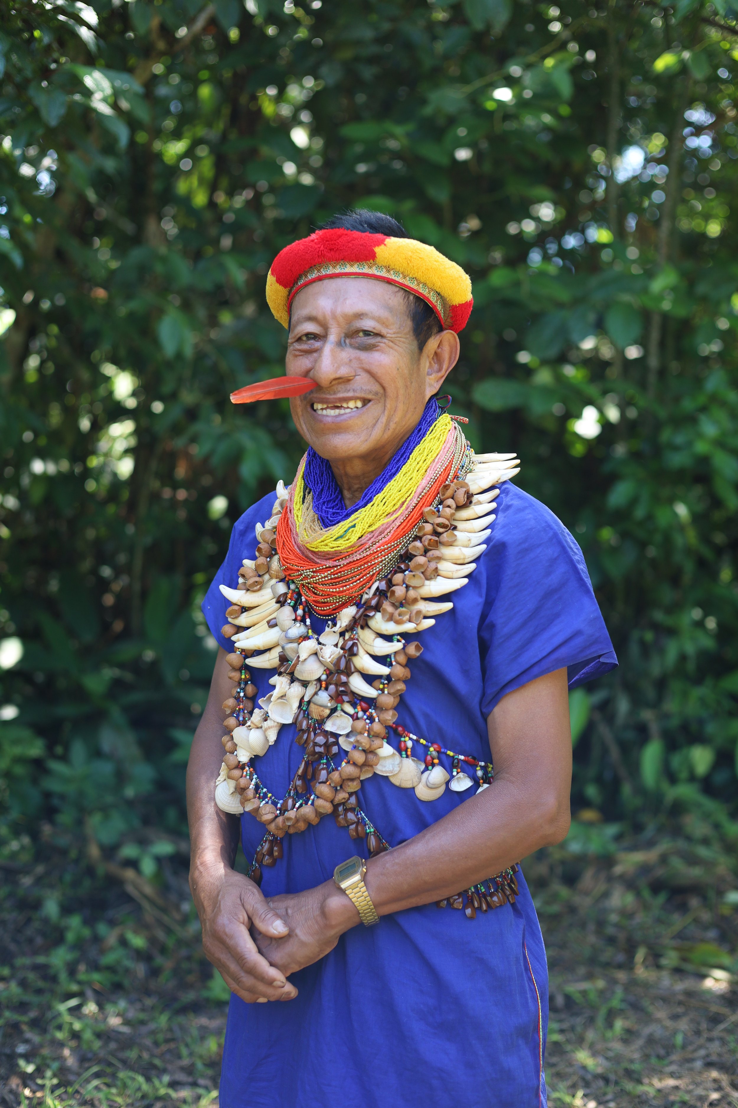 An elderly man wearing traditional tribal jewelry and a colorful hat, standing outdoors with green foliage in the background.