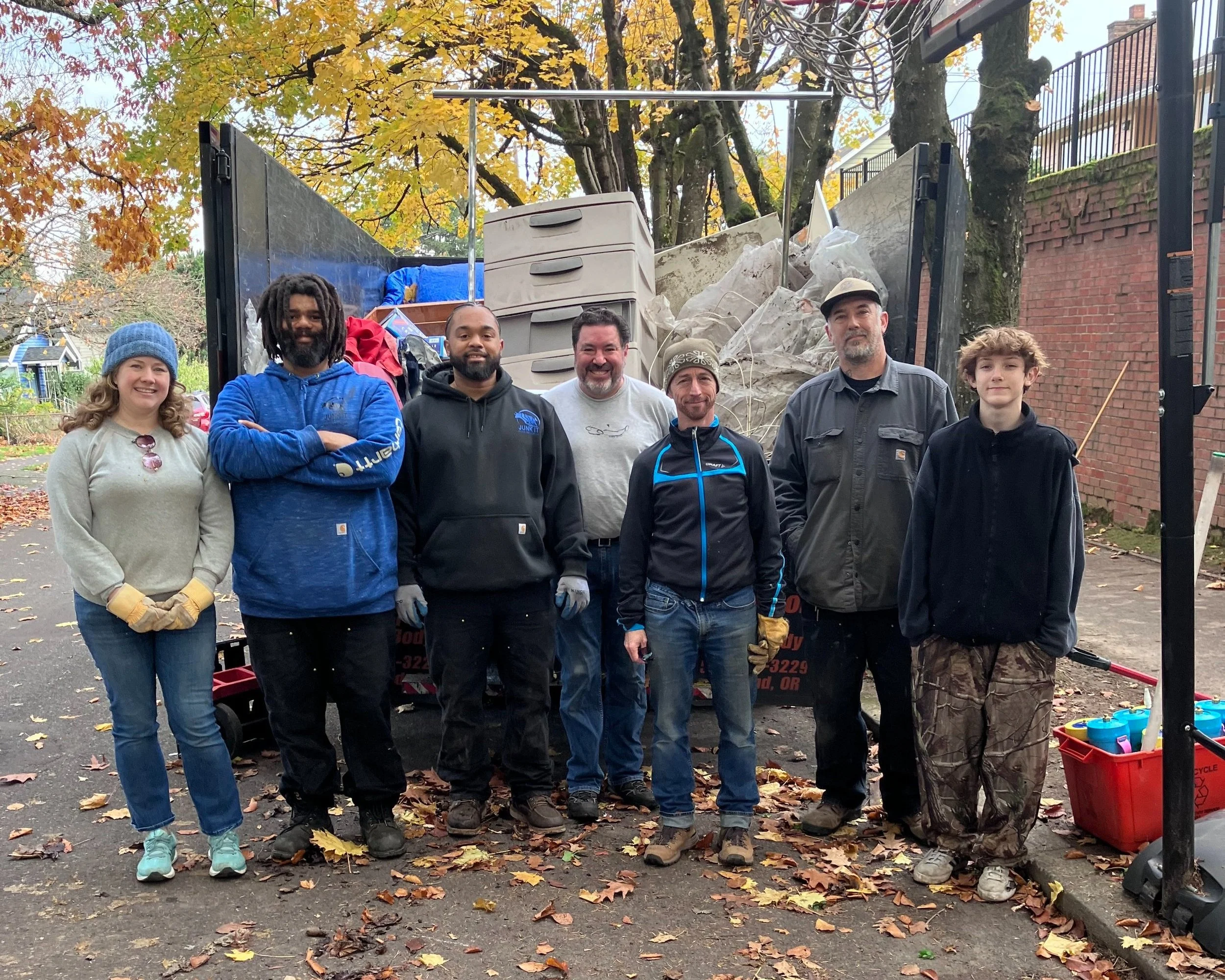 7 Taking Ownership volunteers stand in front of a half-full dumpster on the street during a clean up project.