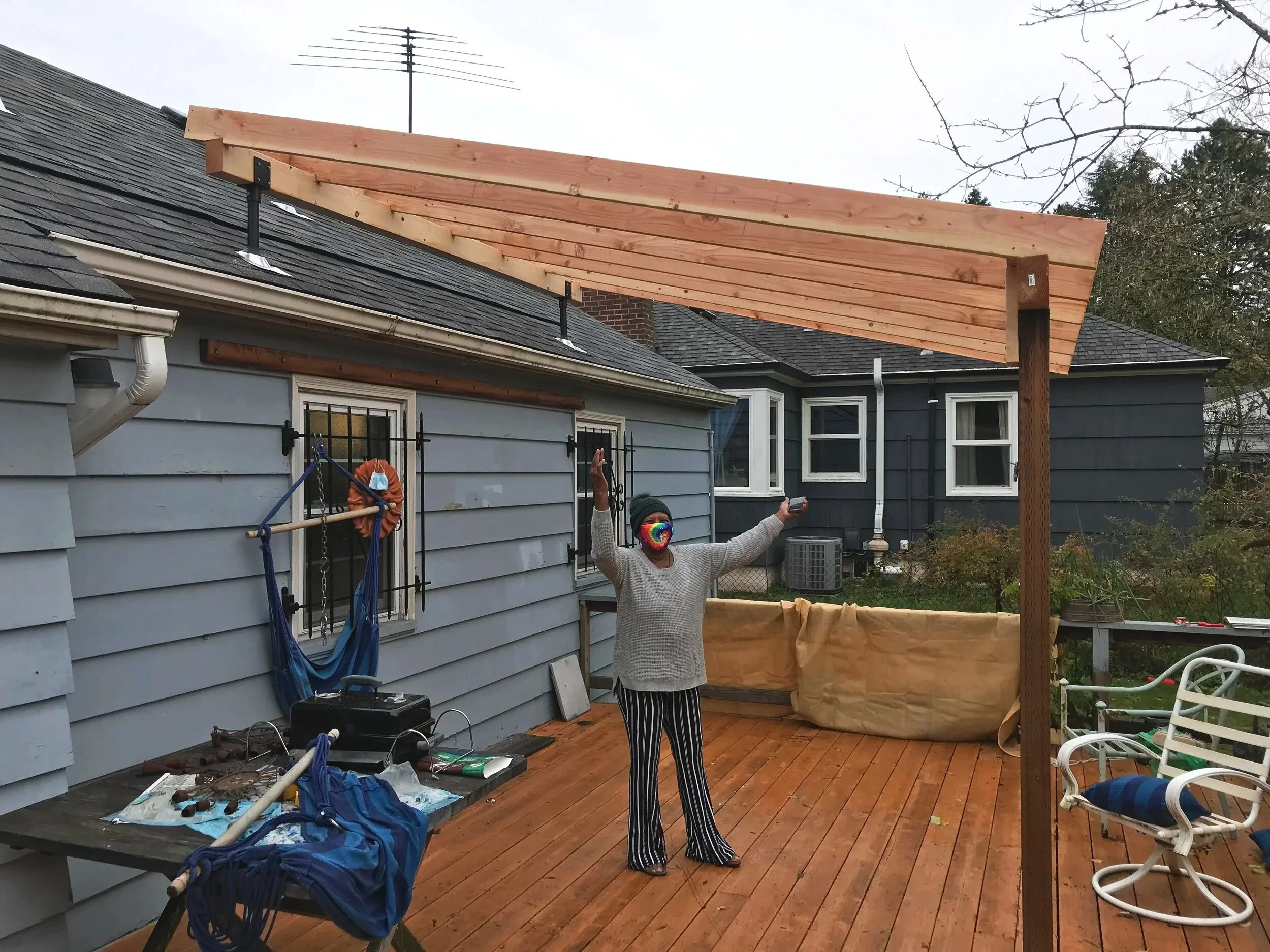 A homeowner wearing a tie-dye mask is holding up her arm underneath a new pergola on her deck.