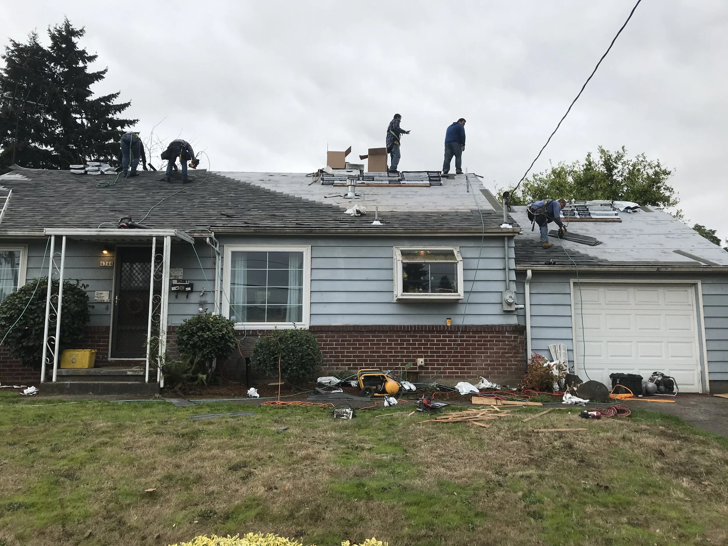 5 people replacing a blue ranch style house roof. Equipment and materials are in front of the house and on the roof where they work.