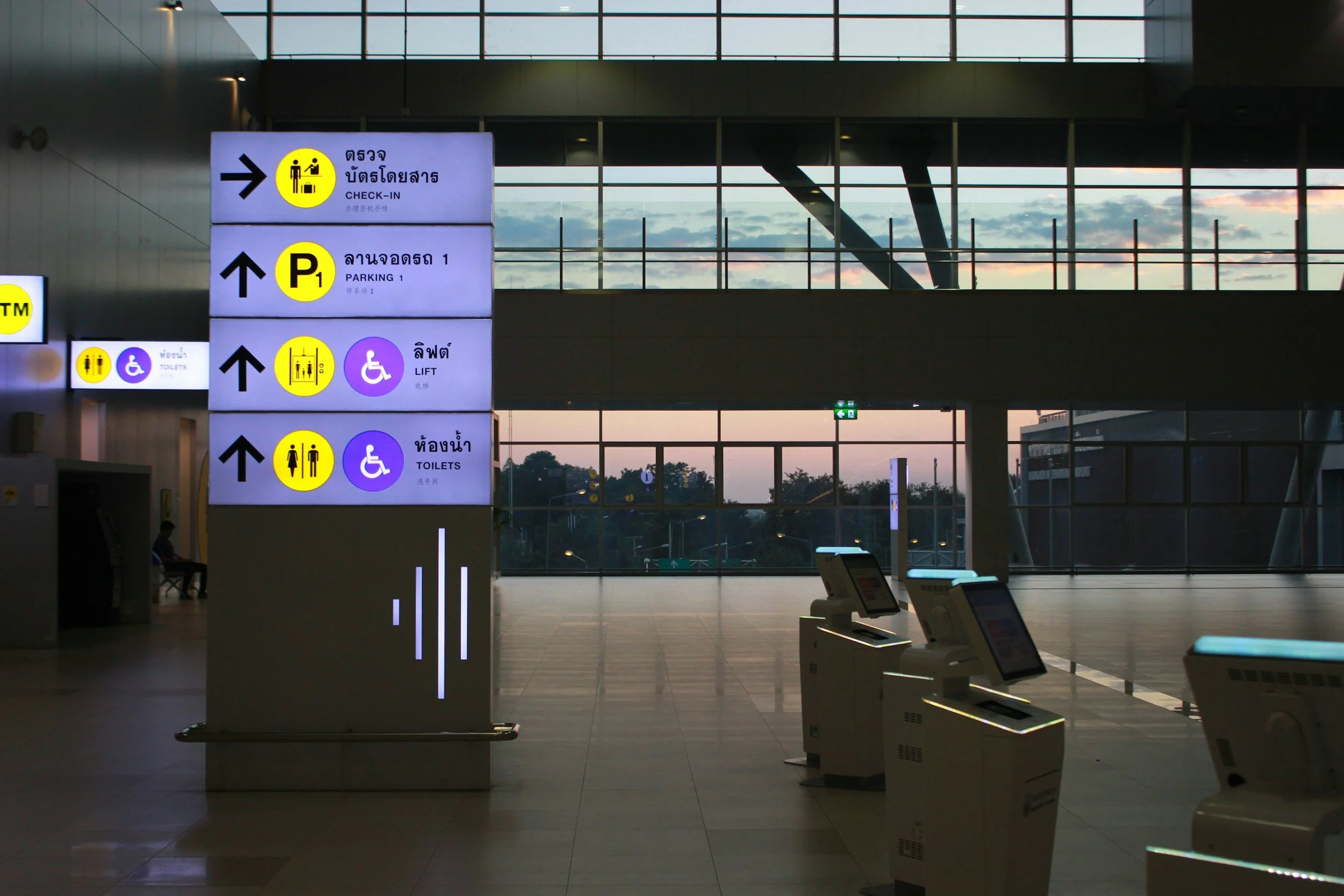 Airport terminal interior with directional signs for check-in, parking, toilets, and lift, seen against large windows showing sunset sky outside.