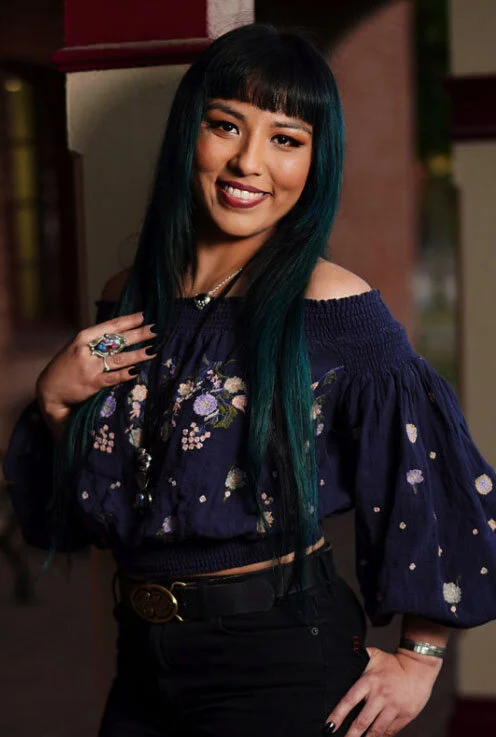 Young woman with long teal hair smiling at the camera, standing indoors. She is wearing an off-the-shoulder navy blue floral top, black pants with a belt, and has rings and a bracelet on her left hand.