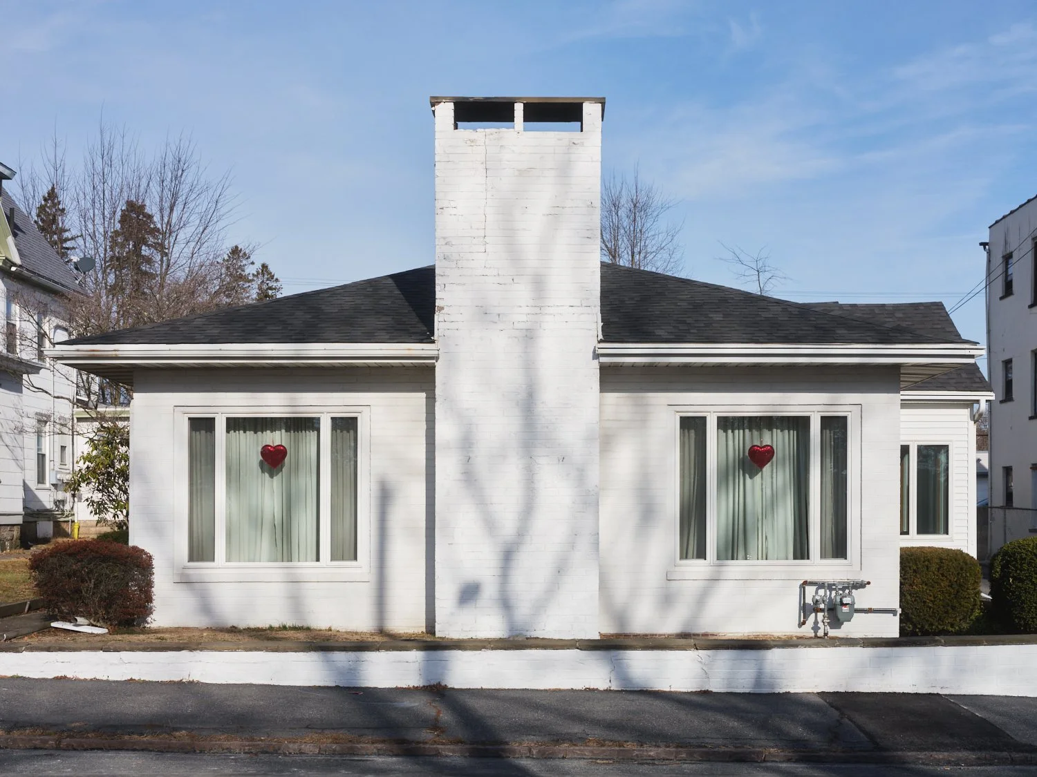 A white house with two large windows, each having a red heart-shaped decoration, and a prominent brick chimney in the center. The house has a dark shingled roof and small bushes on both sides.