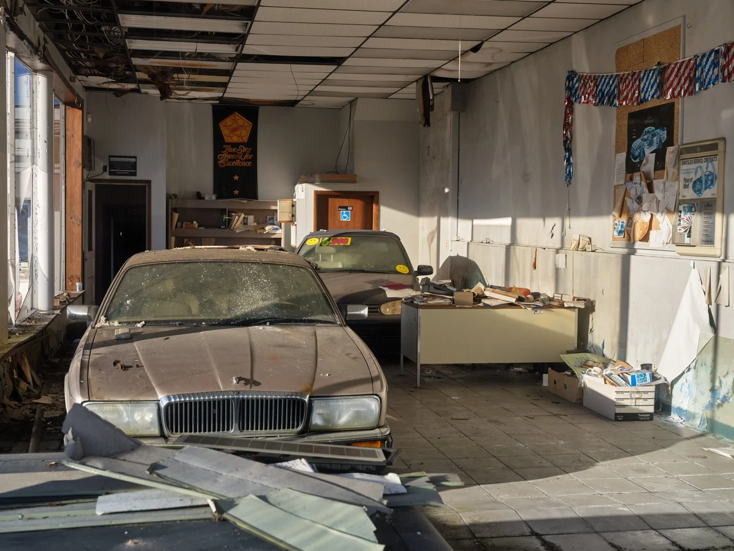Inside a dilapidated, abandoned auto repair shop with two dusty, old cars, debris on the floor, peeling ceiling panels, and cluttered desks and shelves on the wall.