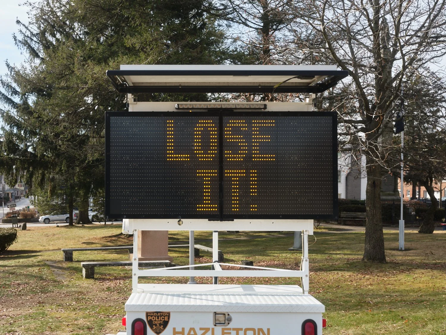 Mobile electronic traffic sign displaying the message "LOSE 7-10" in yellow lights, parked in a park with trees and buildings in the background.