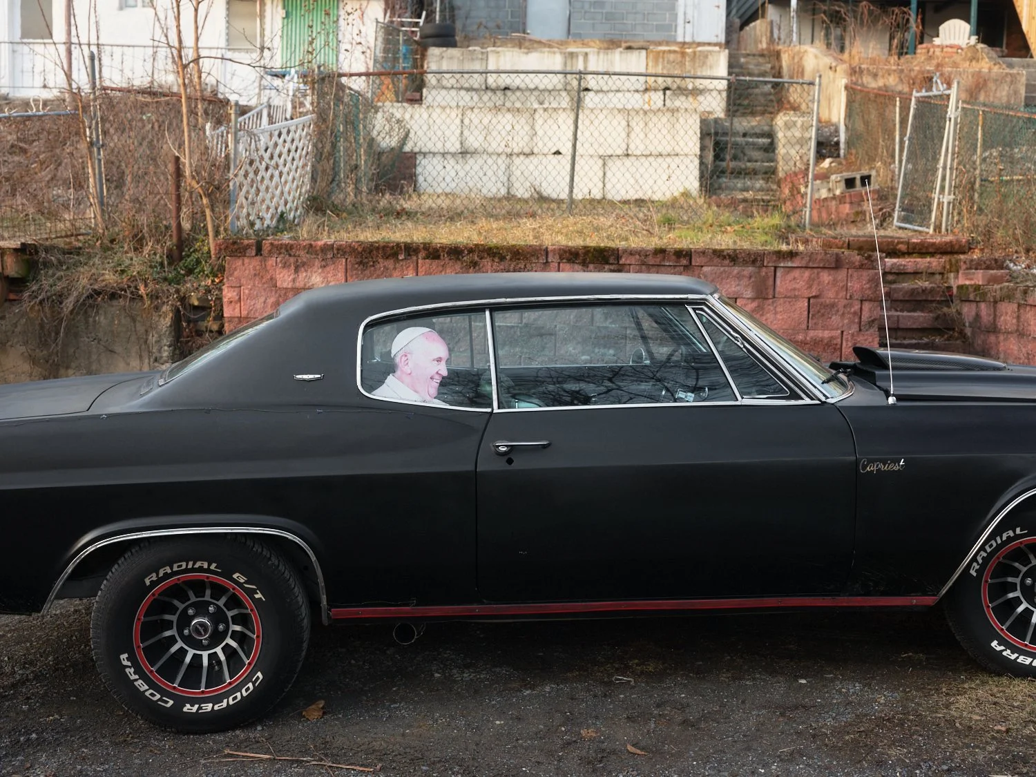A black vintage muscle car parked outdoors with a cardboard cutout of Pope Francis in the driver’s seat. The car has Radial GT tires with red accents and is parked on a gravel surface with a background of brick and wooden fences, stairs, and an overg