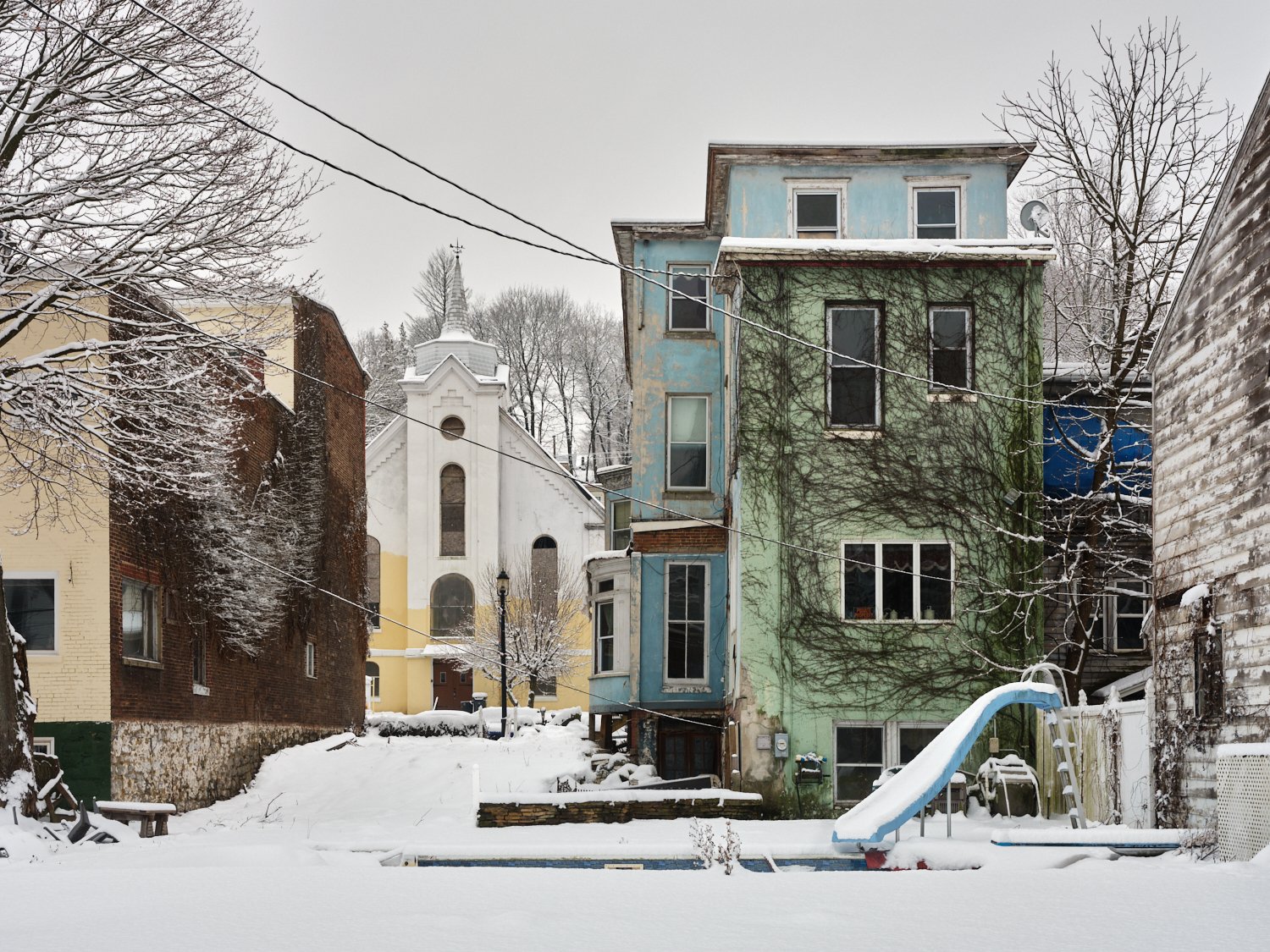 Snow-covered backyard with colorful houses, a playground slide, and a church in the background