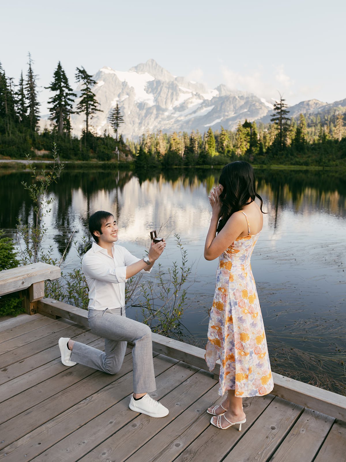 A man on one knee proposing marriage to a woman by a lake with mountains and trees in the background.