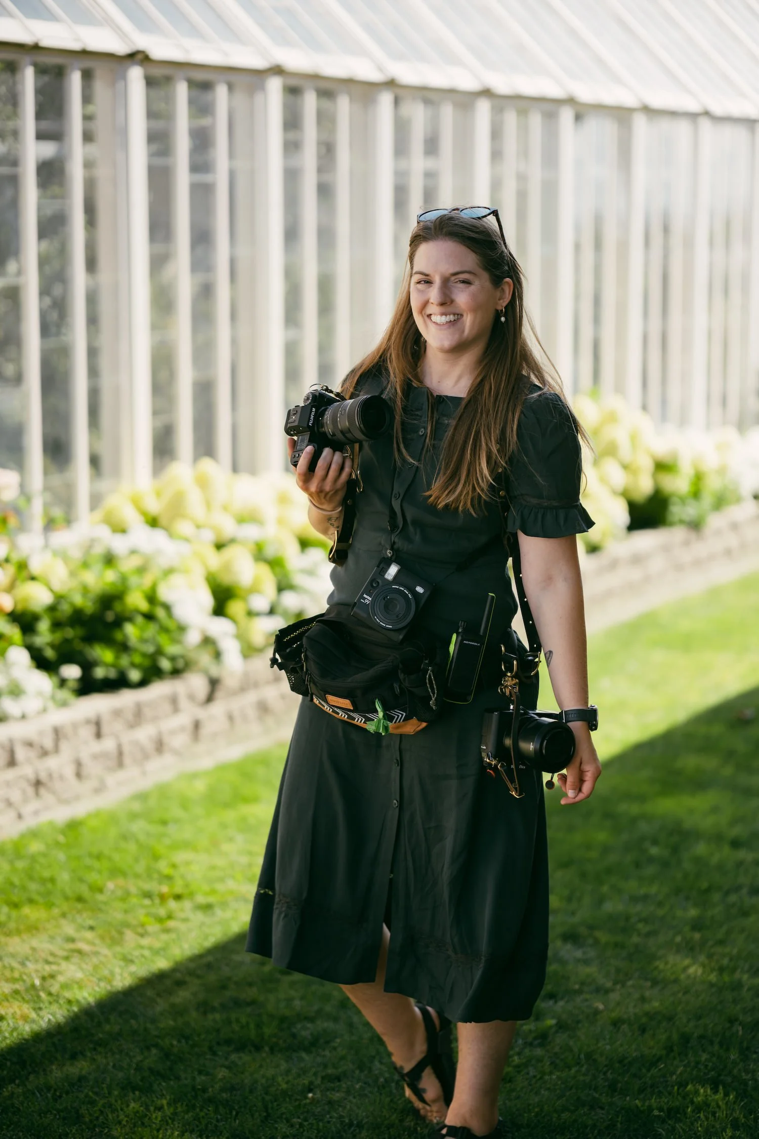 A woman in a black dress holding a camera and a tripod, standing outside in a garden greenhouse with green plants and a metal structure roof in the background.
