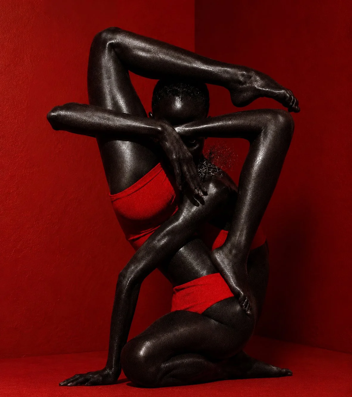 A person with dark skin, dressed in red underwear, poses against a red background with their arms and legs intertwined, creating a complex and artistic posture.