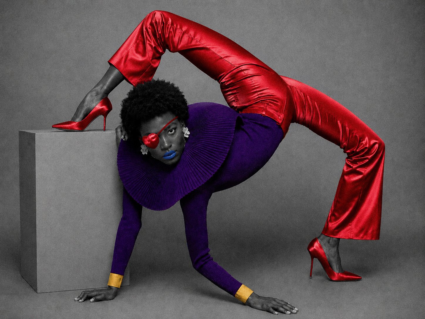 A woman with dark skin, afro hair, and blue lipstick, wearing a purple ruffled collar, purple sweater, and red satin pants. She has a red heart-shaped eyepatch over her right eye, yellow cuffs on her wrists, and red high heels. She is in an artistic 