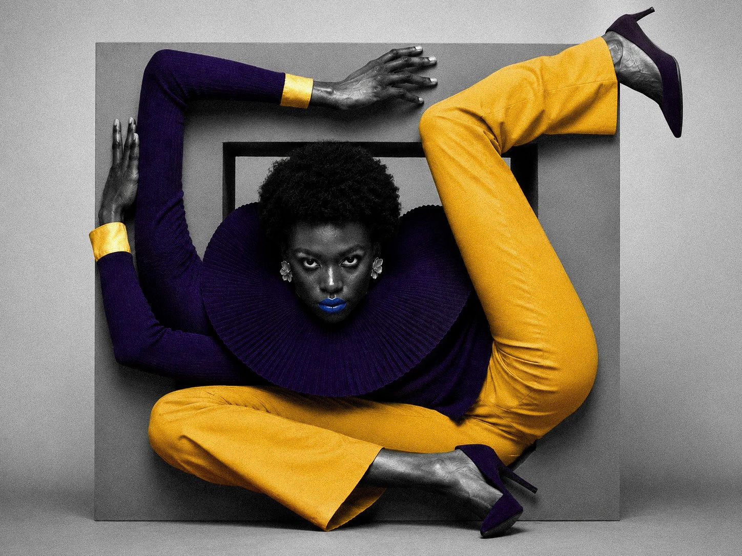 A woman with dark skin and short curly hair poses in an artistic, fashion-forward photo. She is wearing a purple top with ruffled collar details, yellow pants, and purple high heels. She has bright blue lipstick and floral earrings. She is positioned