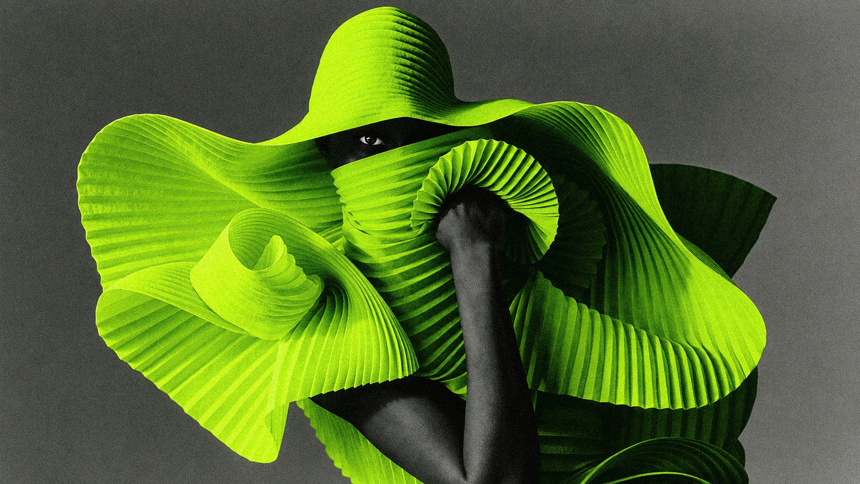 A person with a partially obscured face, covered by large, vibrant green, pleated leaves, with only one eye visible, against a dark background.