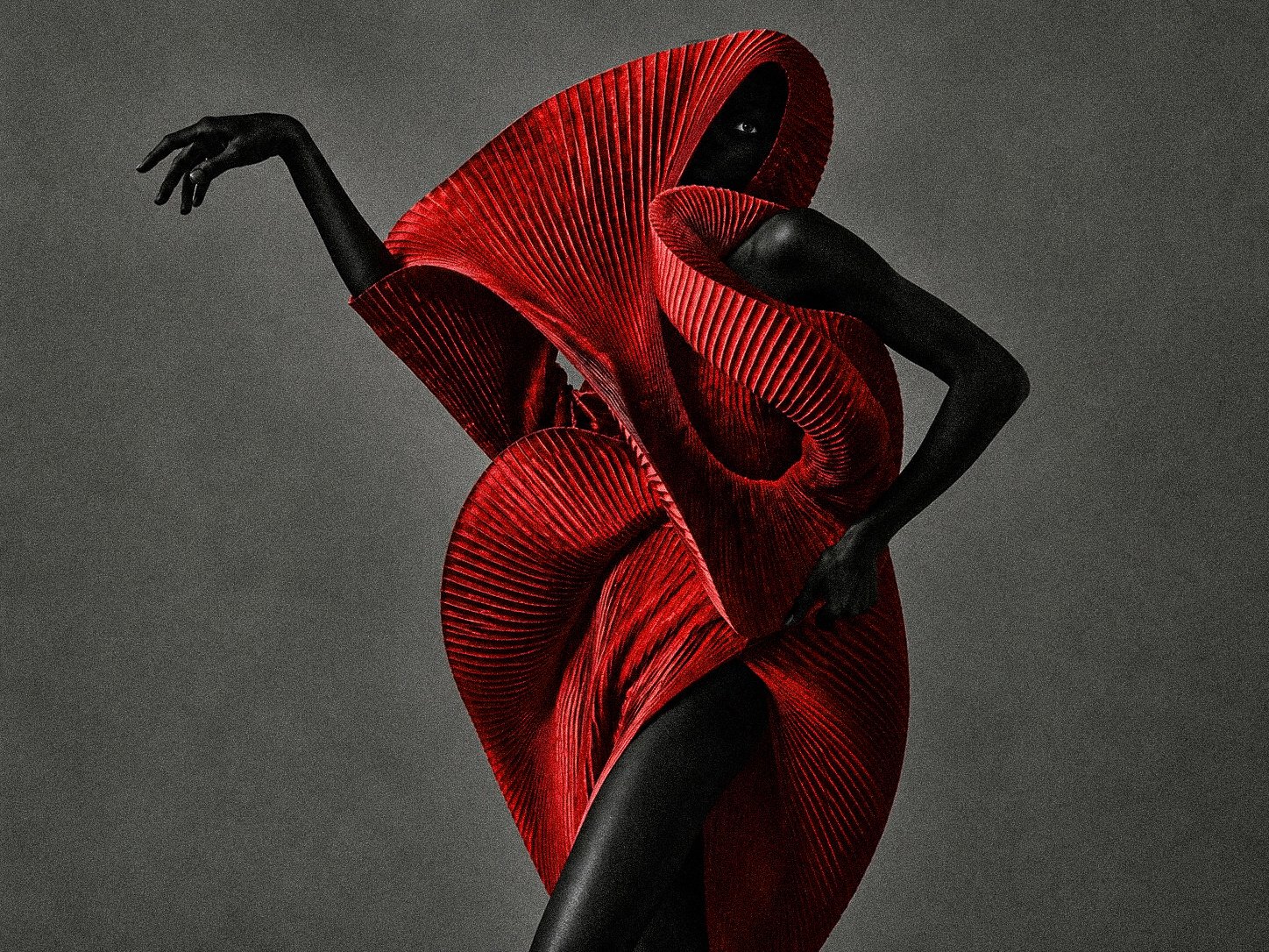 A model wearing an avant-garde, structured red dress with sculptural, pleated details, standing against a dark background.