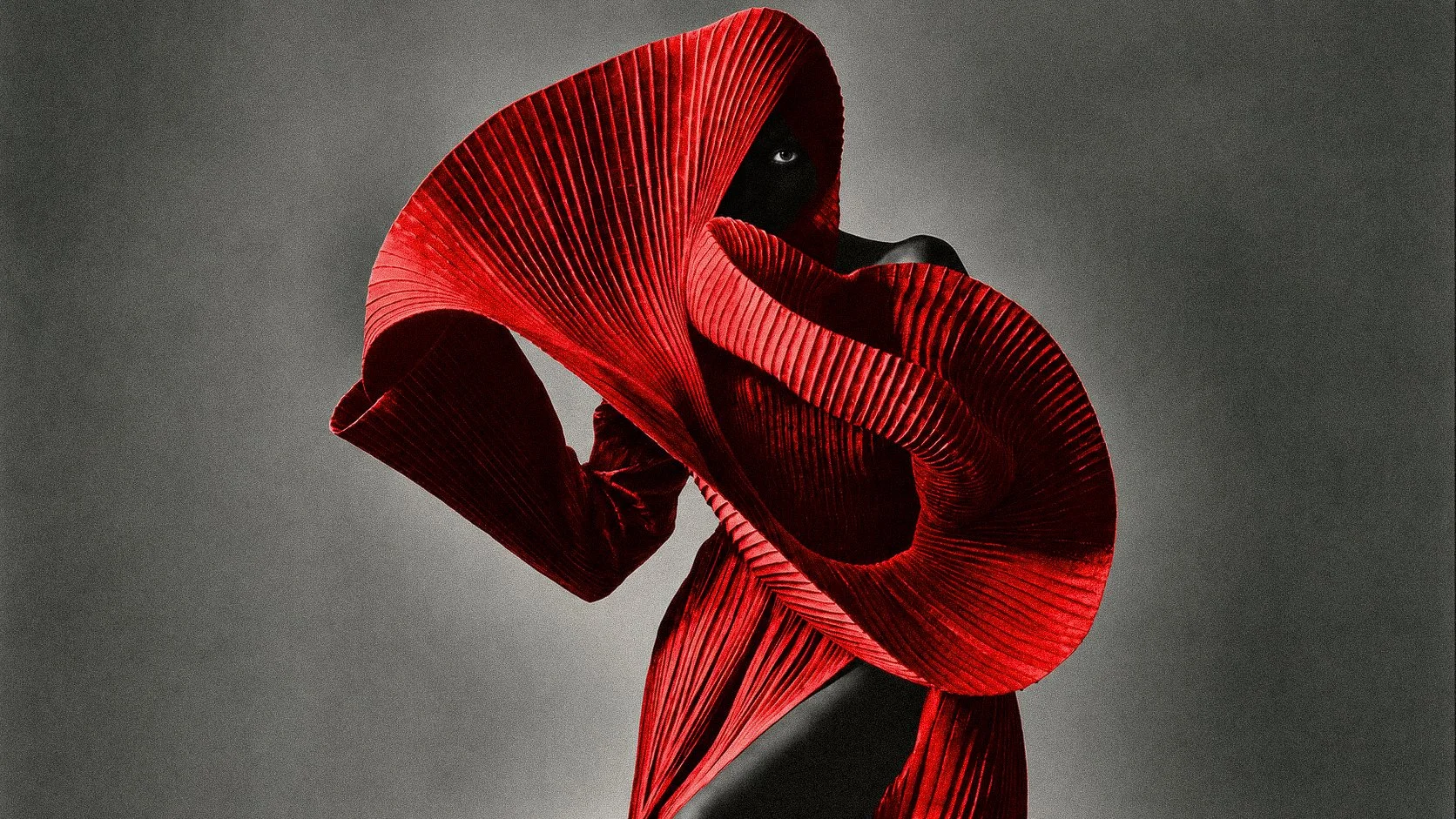 A person with black skin wearing an avant-garde red garment with large, structured, pleated, and curved fabric pieces, standing against a dark gray background.