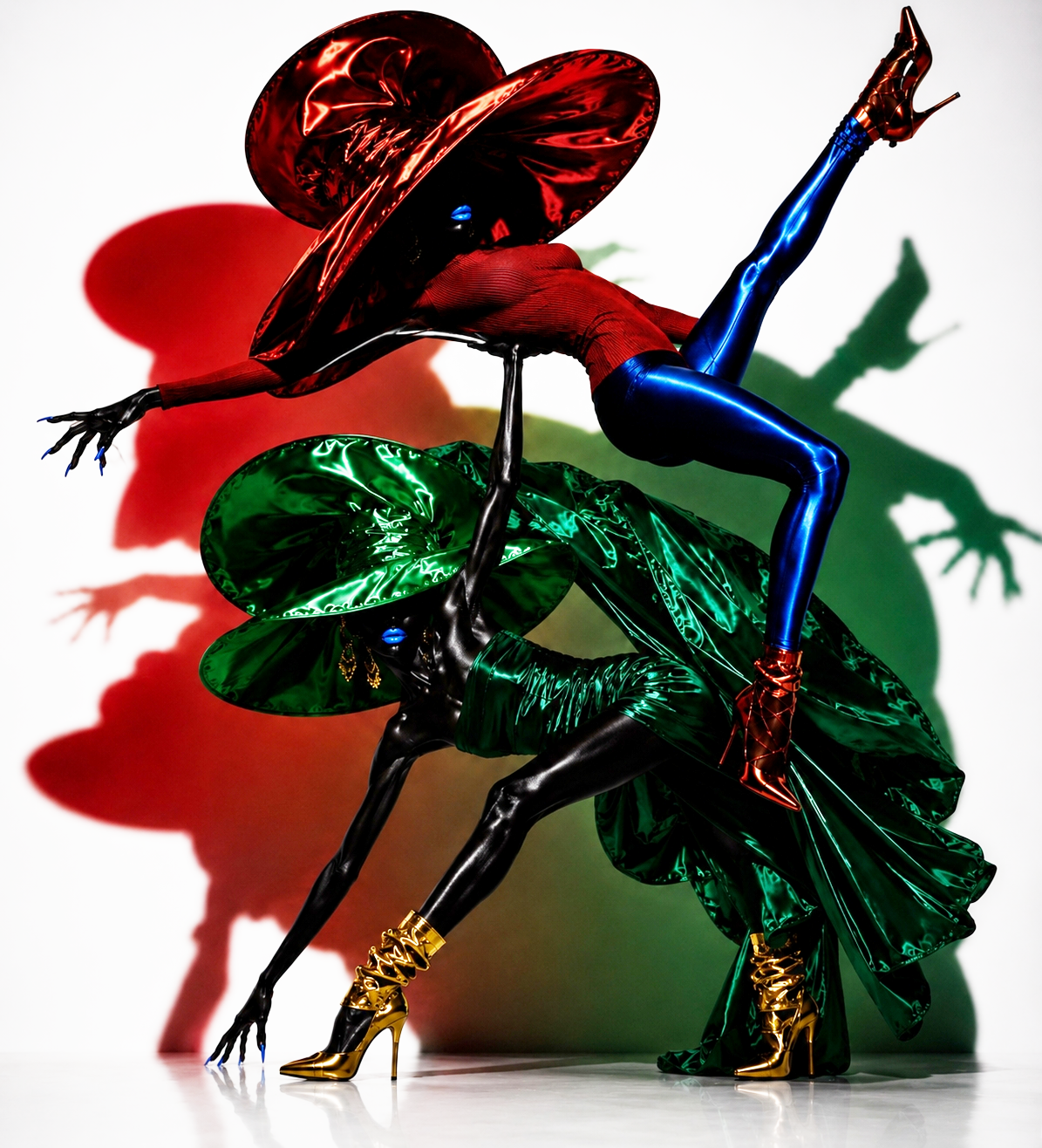Two mannequin figures dressed in colorful, shiny clothing with large green and red hats, posed against a white background with shadows cast in matching colors.
