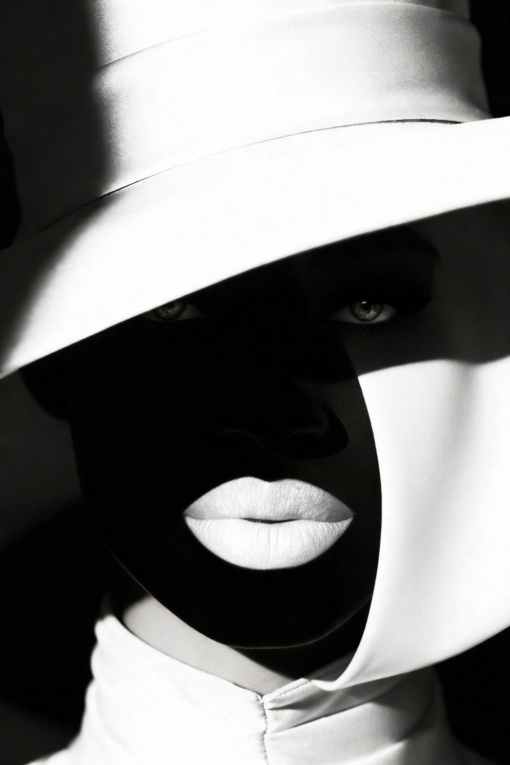 Close-up black and white photo of a woman wearing a wide-brimmed hat that casts a shadow over her face, with only her lips and eyes visible.