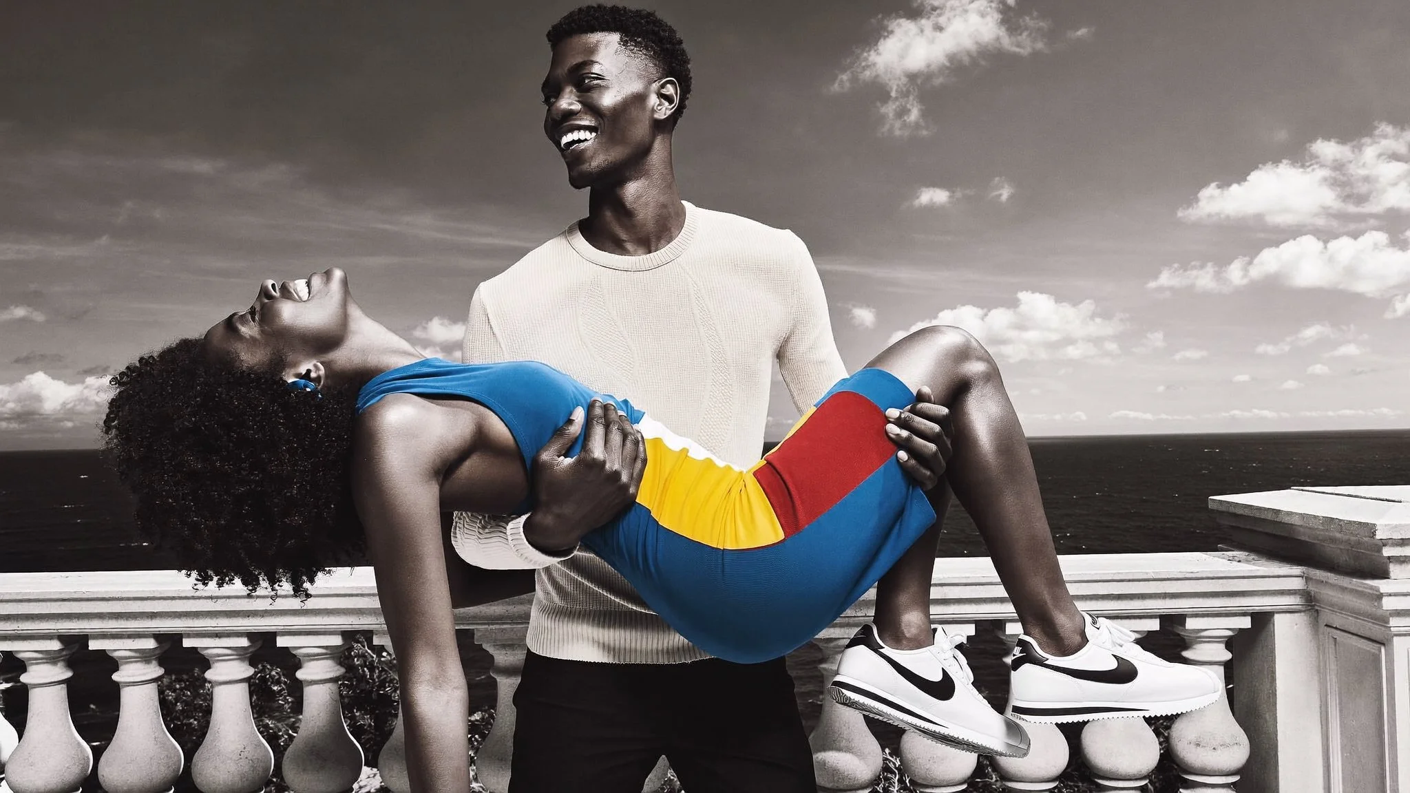 A man holding a woman in his arms outdoors by the ocean, with a cloudy sky in the background. The woman is leaning back with her eyes closed, and the man is smiling while looking at her. The image has a mix of black-and-white and color, with the woma