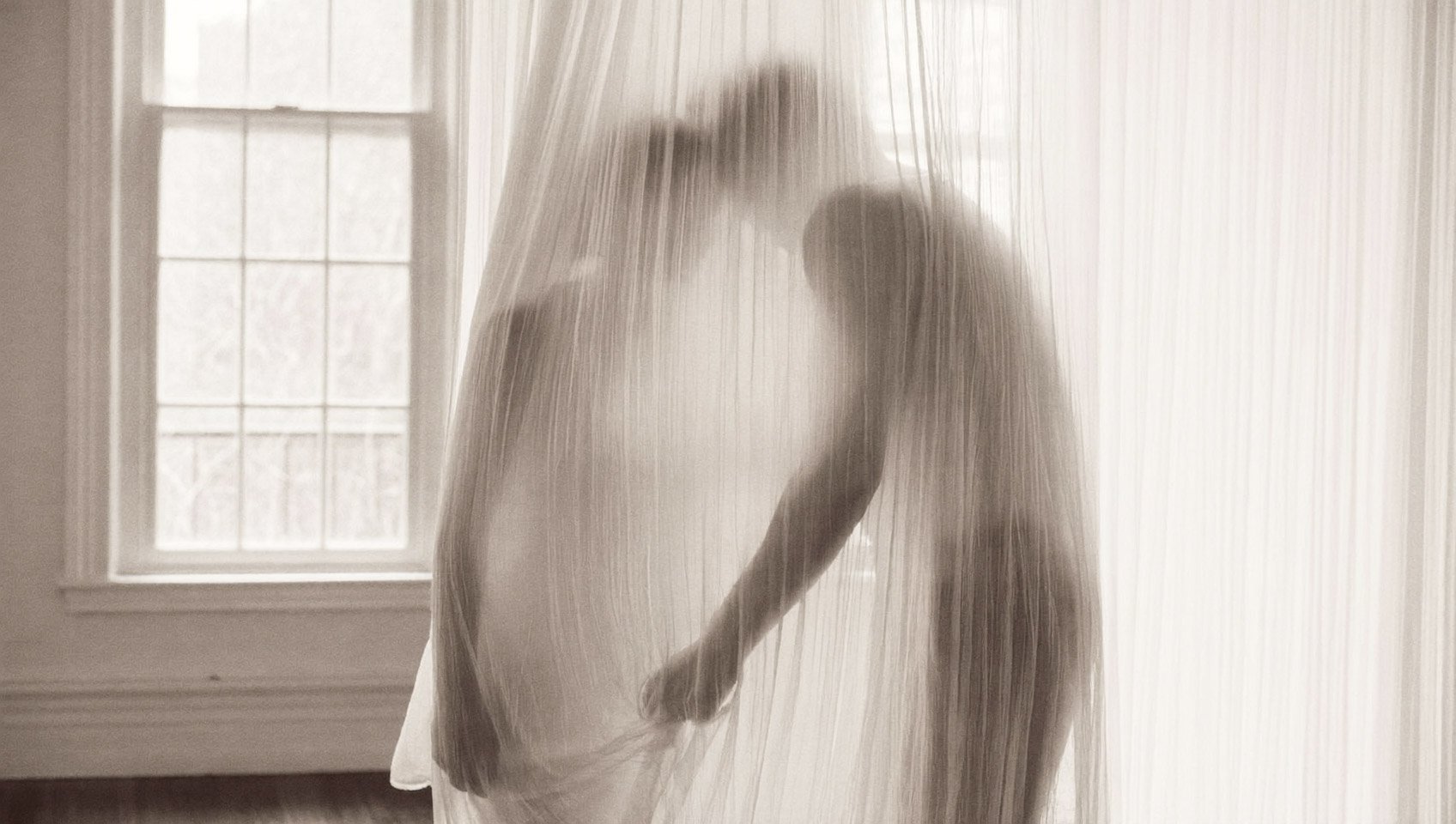 A person stands behind a thin, semi-transparent curtain with a window in the background, softly lit with natural light.
