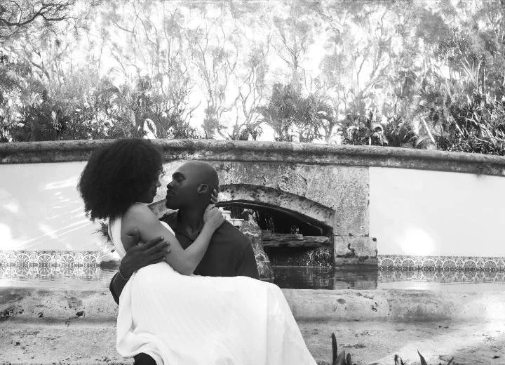 A black and white photo of a man holding a woman in his arms, sitting outdoors in front of a stone fireplace and lush trees.