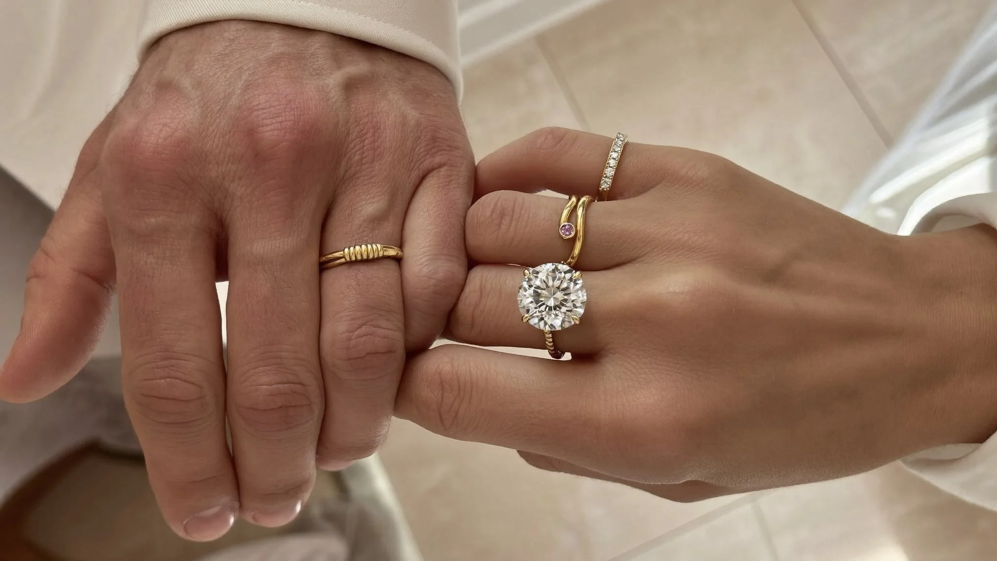 Close-up of two hands intertwined, both wearing multiple gold rings, including one with a large round diamond and others with smaller gemstones.