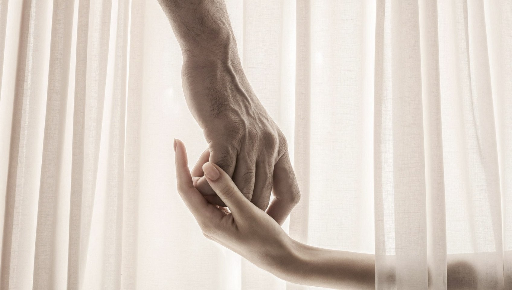 Close-up of a small hand holding the finger of a larger hand in front of a light-colored curtain.