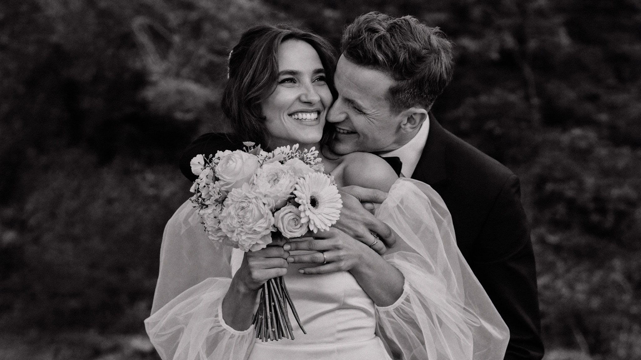 A black and white photo of a joyful couple embracing, with the woman holding a bouquet of flowers. The woman is smiling happily and the man is close to her face, also smiling.