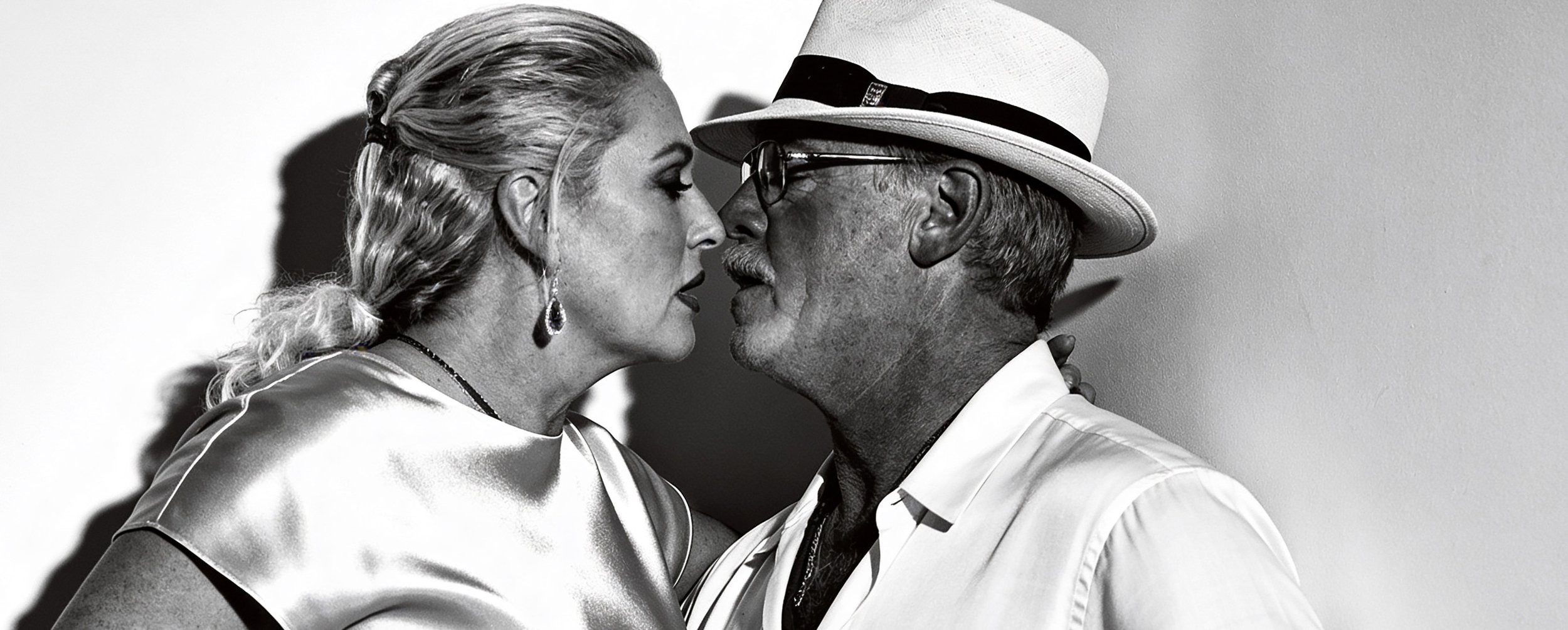A black-and-white photo of a woman and an older man with a fedora hat, standing close face-to-face, about to kiss.