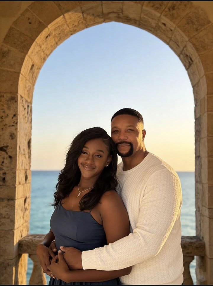 A smiling couple stands close together in front of a stone archway overlooking the ocean at sunset. The woman has long dark hair and wears a blue dress with a cross necklace, while the man has short dark hair and a goatee, wearing a white sweater.
