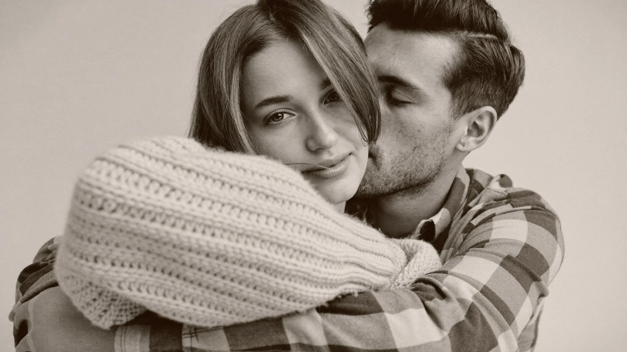 A young woman with long hair and a young man with short hair embracing each other. The woman is looking at the camera with a slight smile, while the man has his eyes closed and is kissing her cheek.