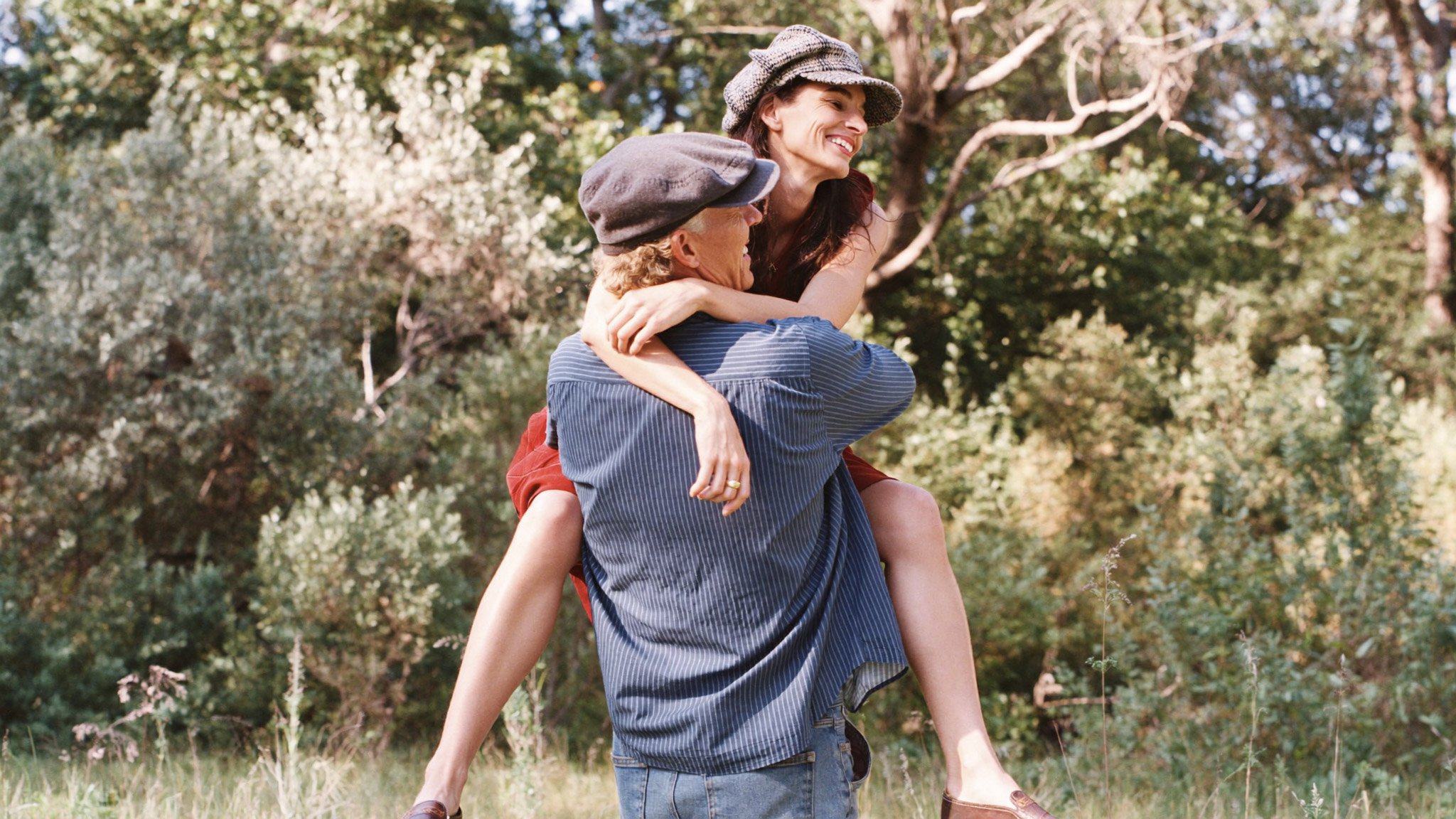 A woman sitting on a man's shoulders outdoors in a park or forest, smiling and enjoying the moment.