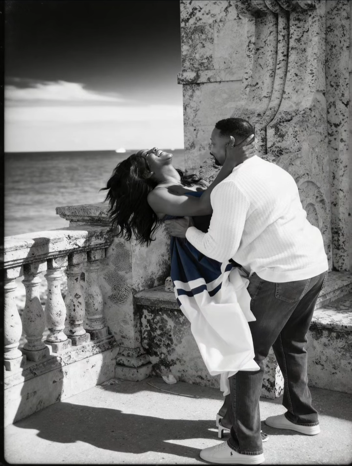 A man and a woman share a romantic moment by the seaside, with the woman leaning back and the man holding her, near an old stone wall and balustrade.