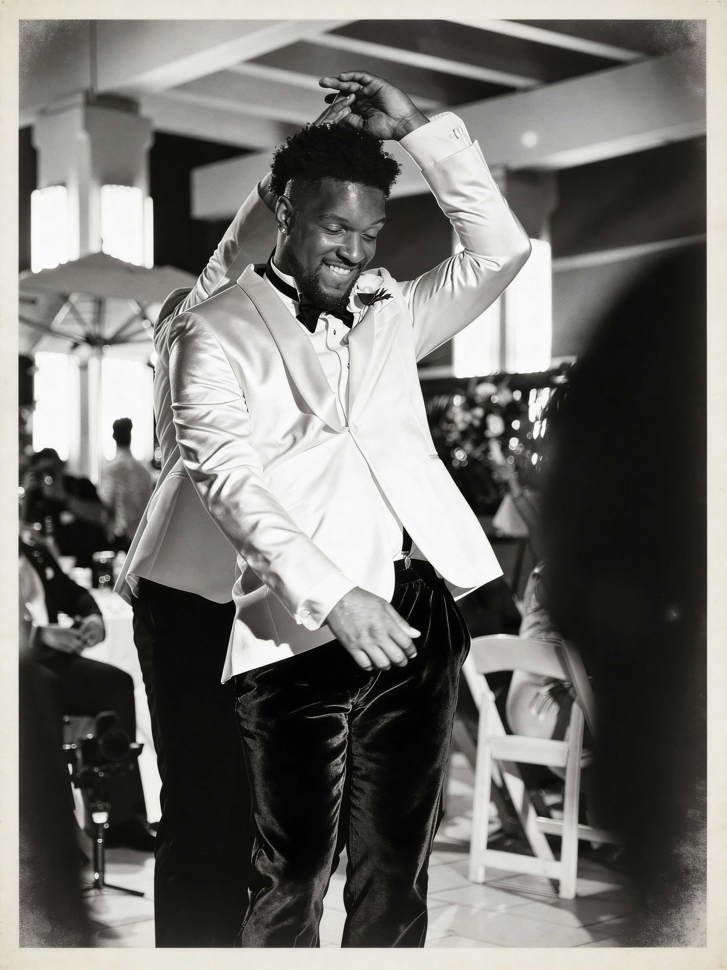 A black-and-white photo of a man dancing, wearing a shiny tuxedo with a bow tie, in a social or dance setting with people seated and standing in the background.