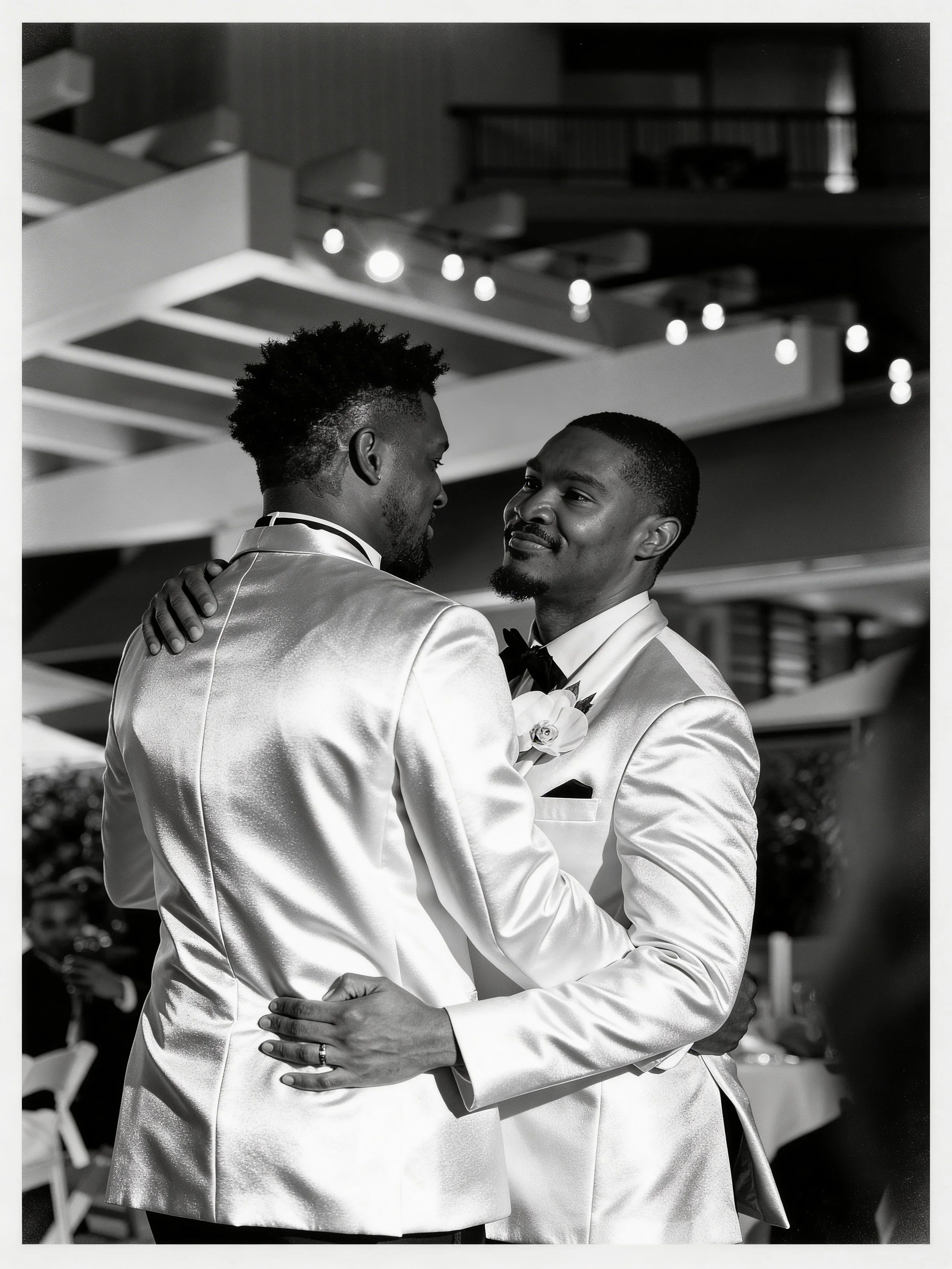 Two men in tuxedos embrace at a formal event, smiling at each other, with a modern indoor setting and decorative lights in the background.