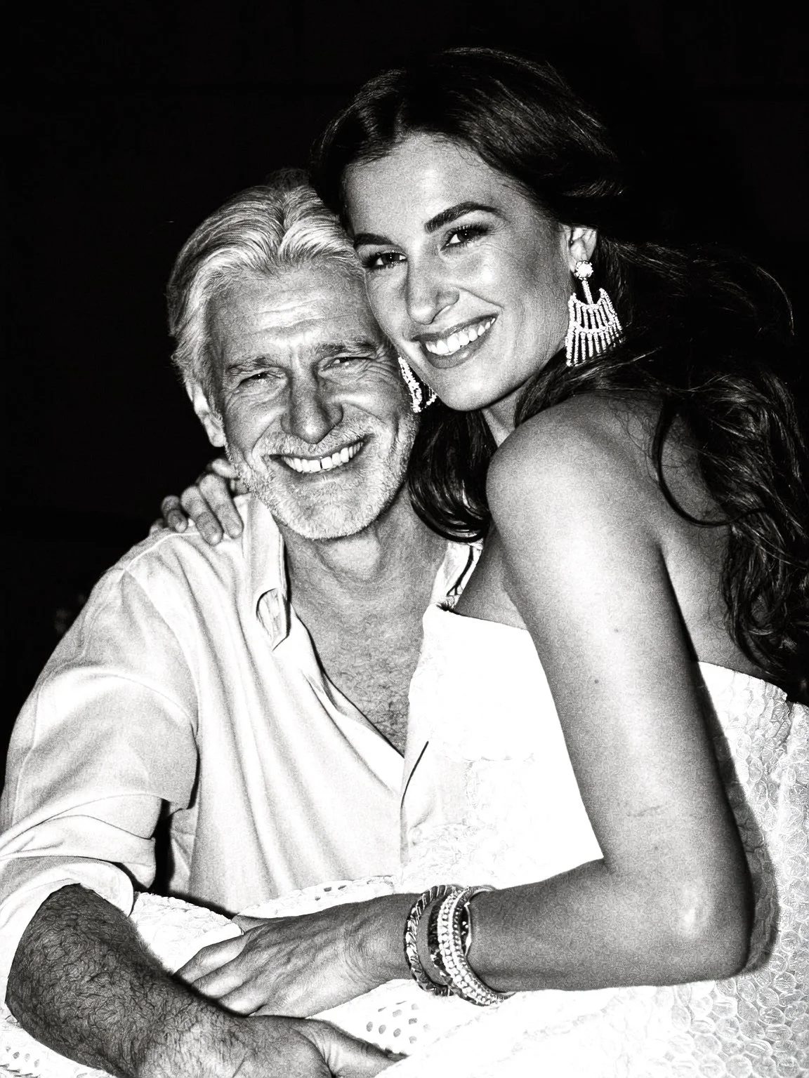 A smiling older man with white hair and a beard, and a smiling young woman with long dark hair, posing close together in black and white.